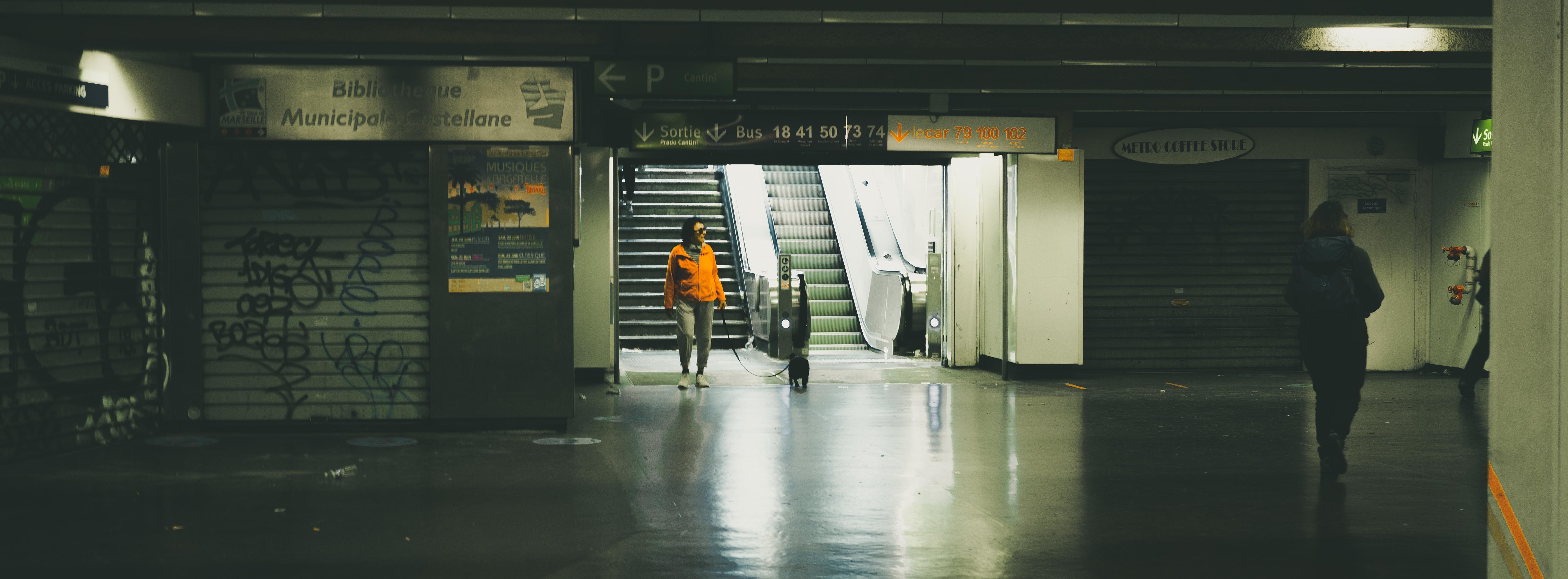 Subway station interior with dim lighting, featuring a solitary figure in an orange jacket walking toward an escalator. The floor reflects the overhead light. Graffiti is visible on a closed shop shutter to the left, and there are signs above the escalator. Another person walks away in the right foreground.