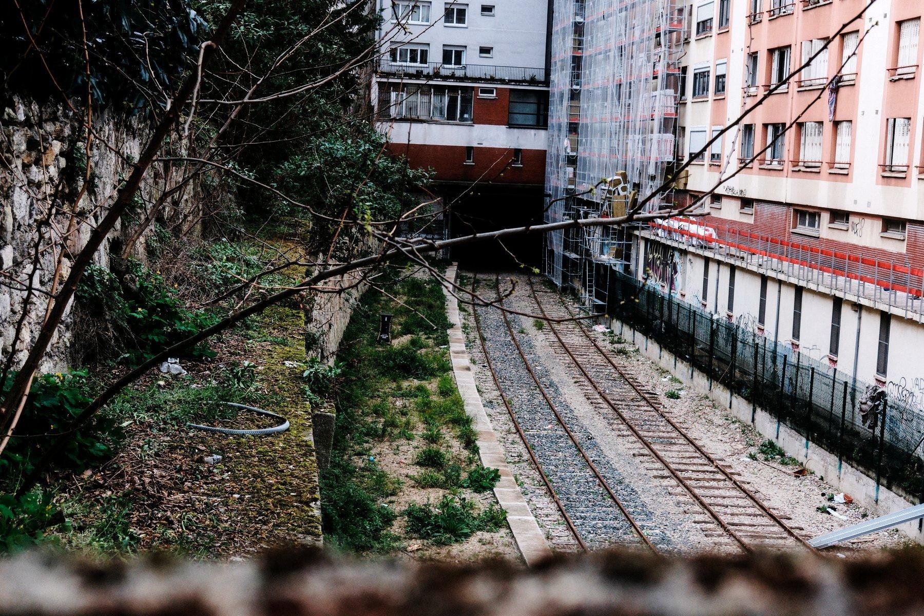 Photo horizontale. Au milieu d’immeuble des années 70-80, deux voies de chemin de fer abandonnées et allant dans un tunnel sombre