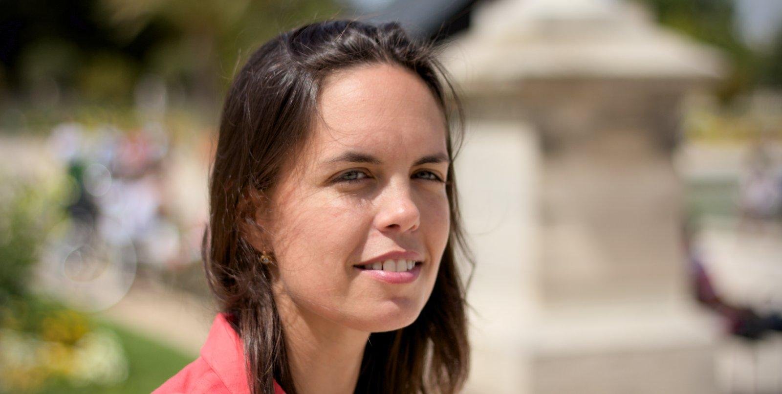 Photo format paysage en 16:9 d’une jeune femme brune aux cheveux long en gros plan. Elle a un grand sourire, éclairée par le soleil. Le fond est totalement flou mais on devine un parc