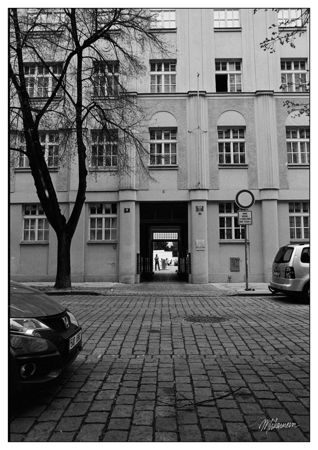 Cobblestone street leads to the entrance of a multi-story building with multiple square windows. Bare trees line the street, and several parked cars are visible. A person stands in the distance through the entrance, and a 'no entry' sign is on the right side near the building.