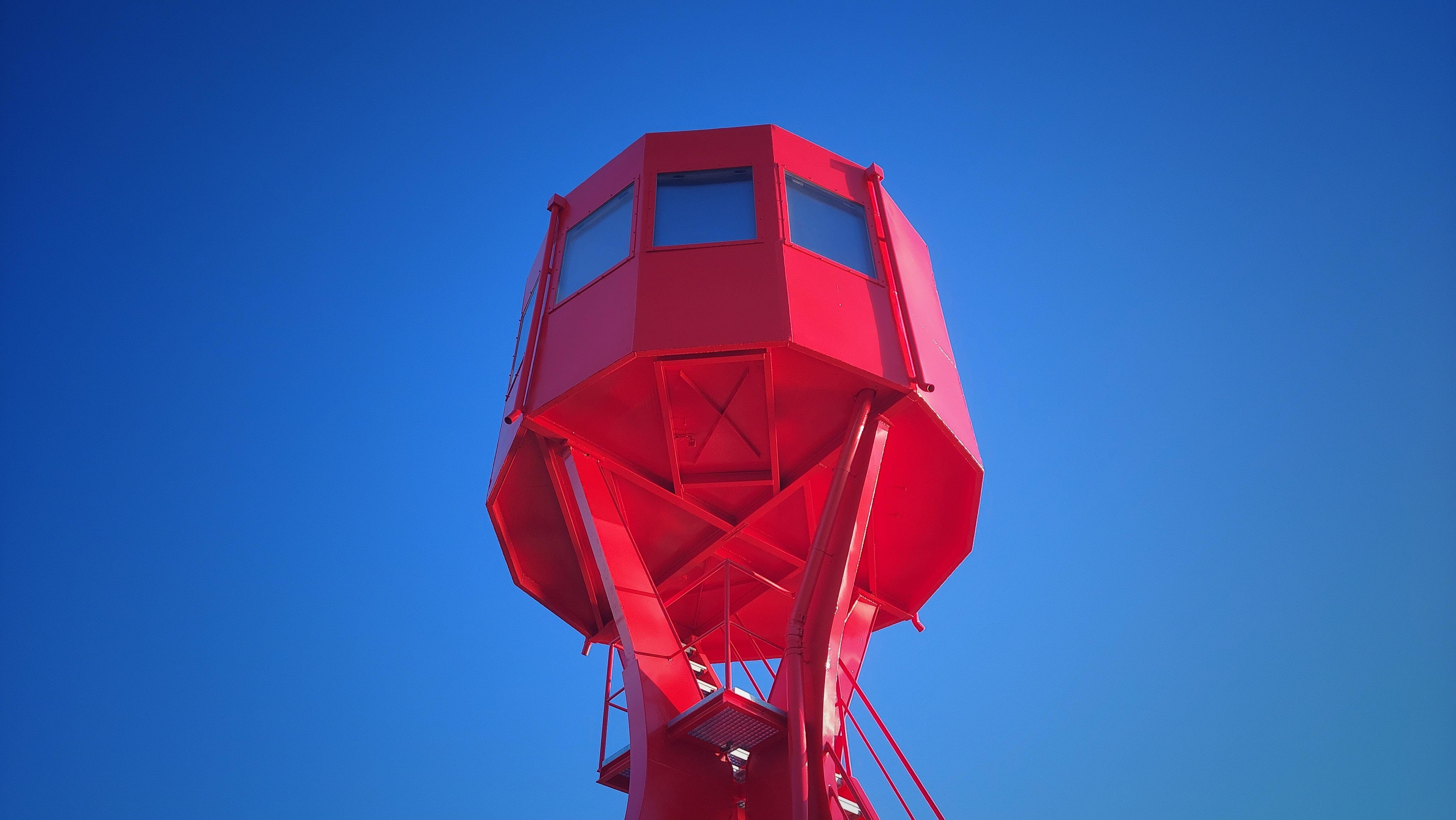 The image shows a red observation tower against a clear blue sky. The tower has a geometric, angular design with windows near the top.