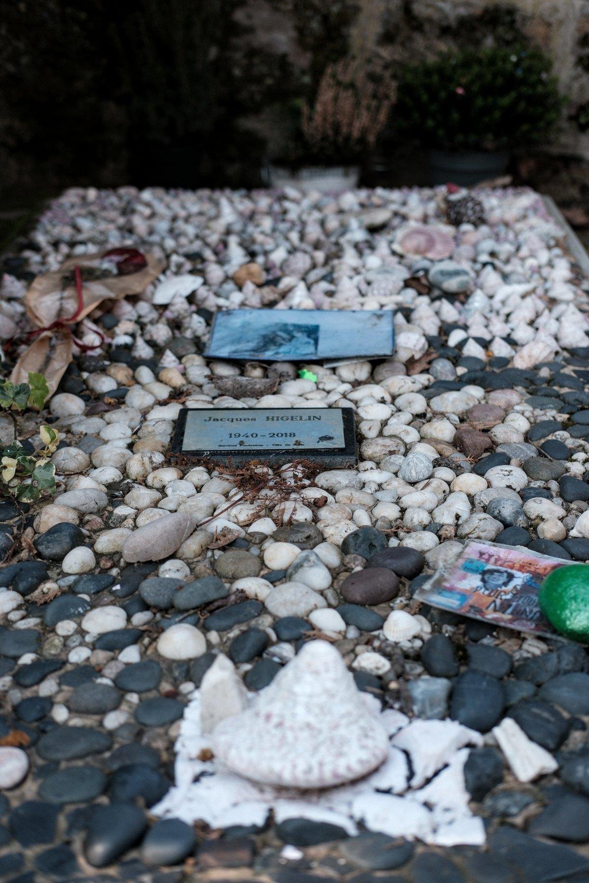 Photo verticale. Une pierre tombale sans stèle, couverte de coquillages et de galets. On peut lire une plaque : Jacques Higelin 1940-2018