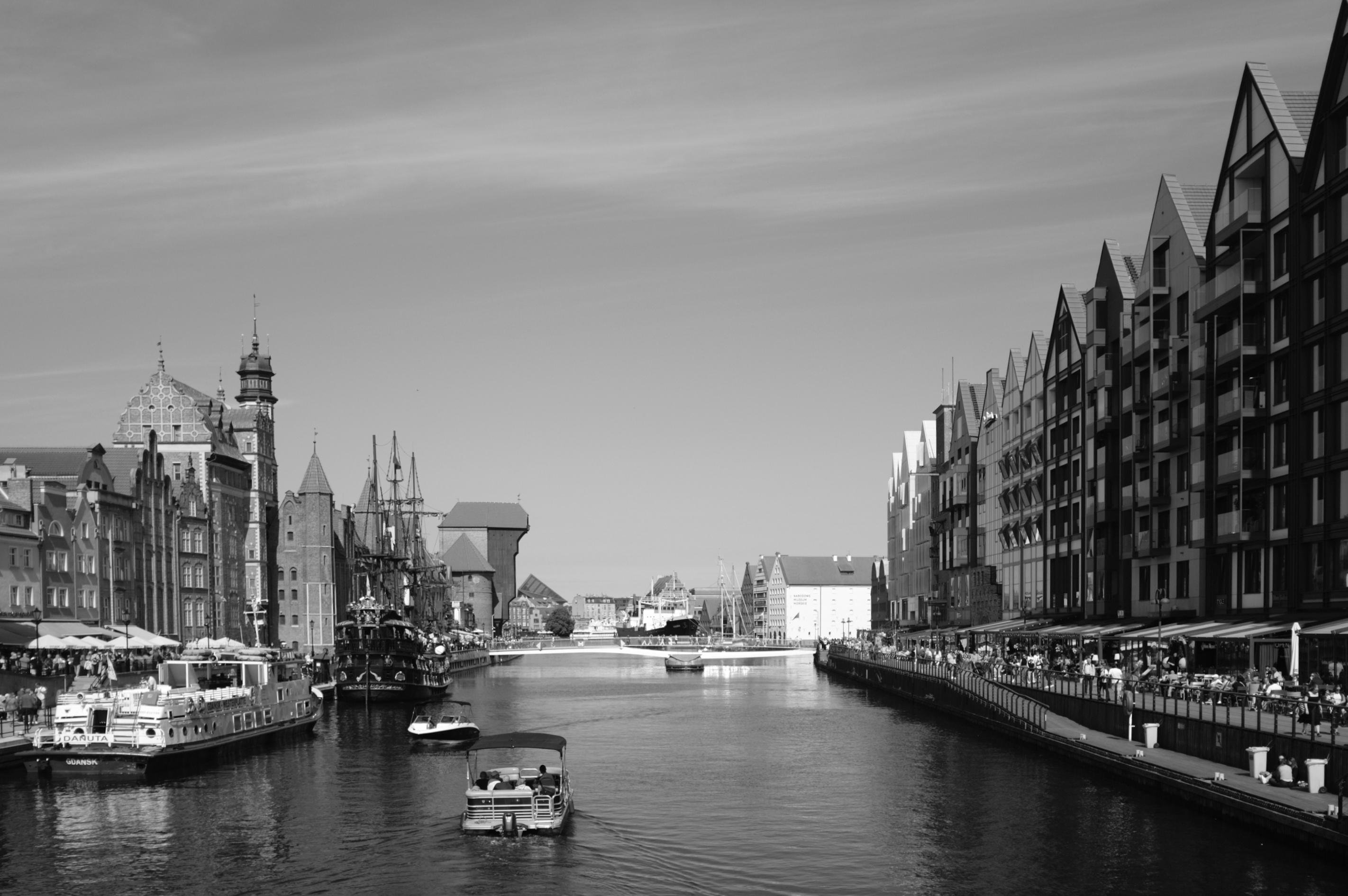 Black and white photograph of a canal flanked by historic buildings on both sides. A small boat moves through the center of the waterway. On the left, a tall clock tower and a line of medieval-style structures, while modern, multi-story buildings with peaked roofs line the right. Sidewalks beside the canal are crowded with people and outdoor seating. A clear sky overhead.