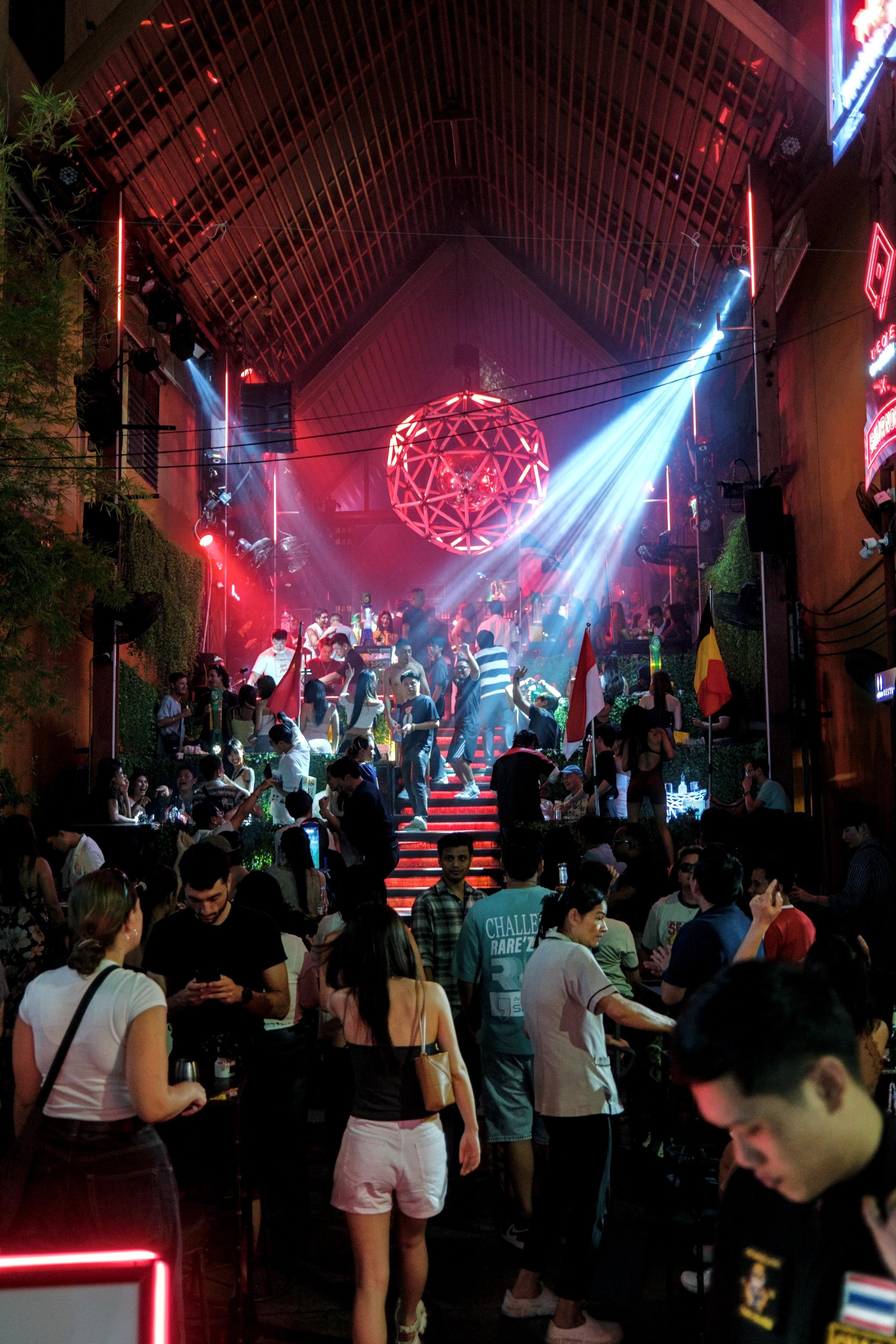 Portrait picture of a crowded open-air bar with people standing and sitting around a wide staircase, red step lights leading up to dance floor placed under a huge red sphere light fixture. White spotlights cut across the scene.