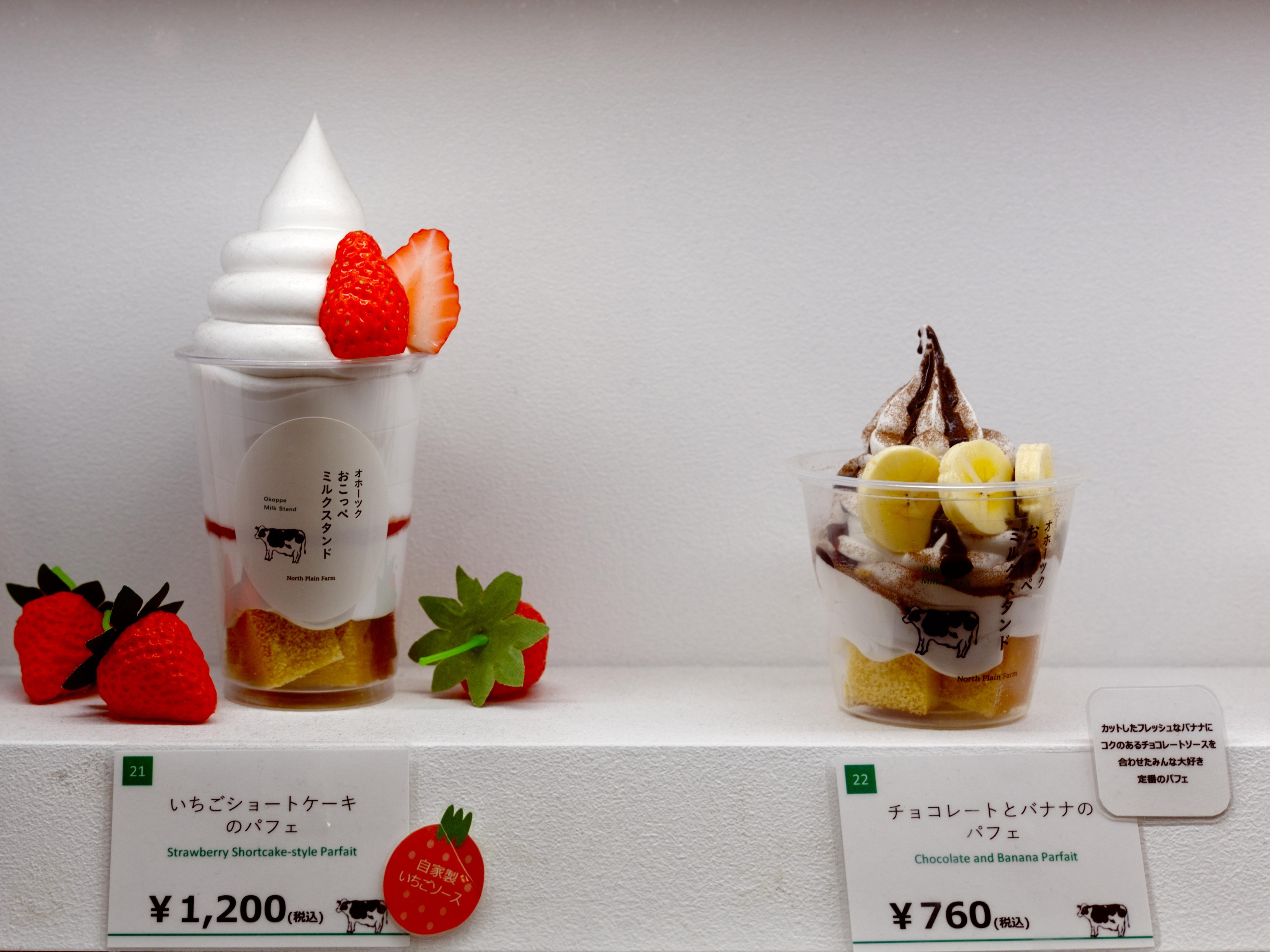 A picture of two parfaits lying against a pure white background with neutral lighting, as if presented as luxury items. The vivid red of the left, strawberry flavored parfait contrasts with the darker chocolate and banana parfait on the right. Little signs describe each item and their price in Japanese and English.