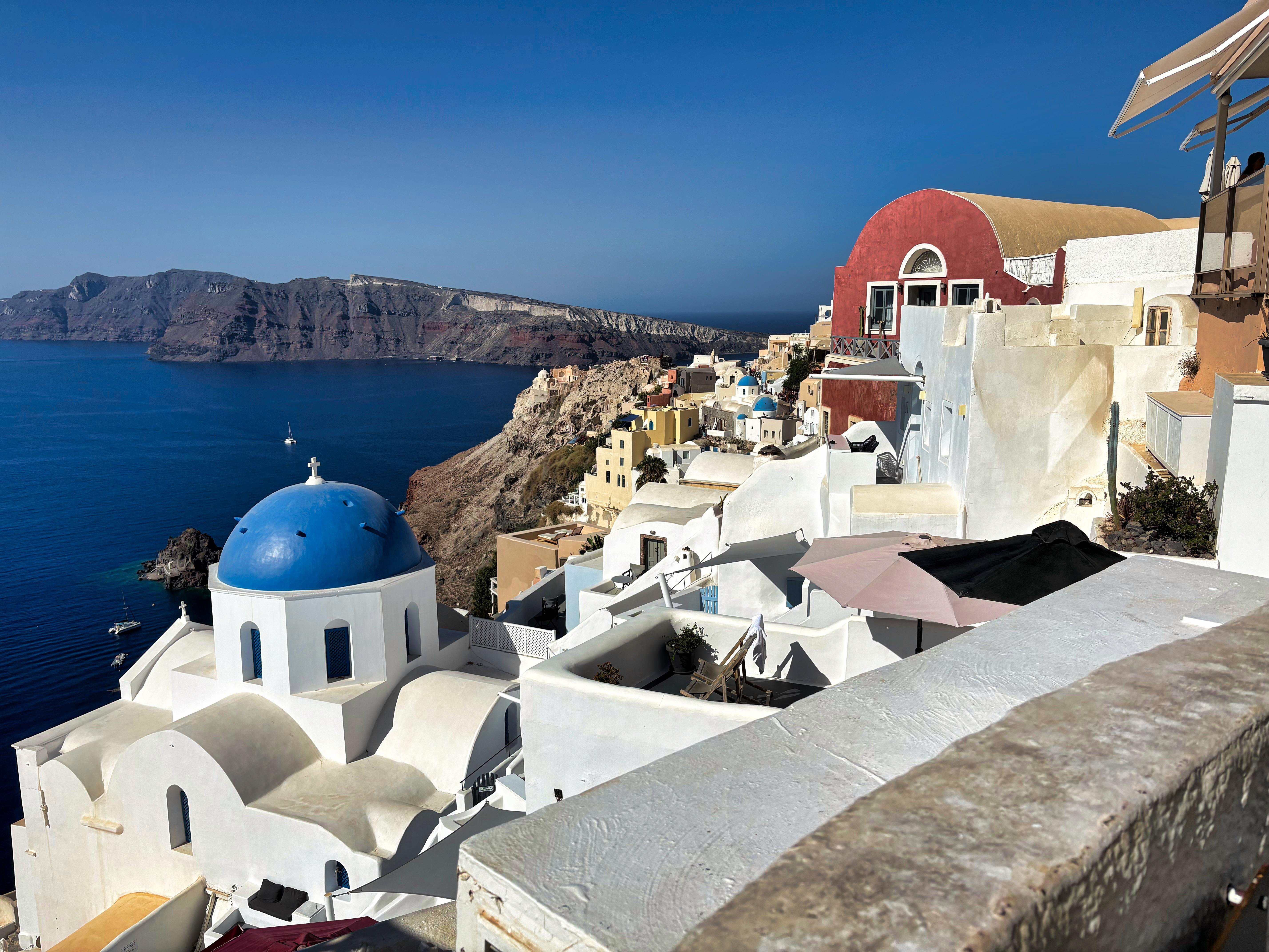 White buildings with blue domes overlook a cliffside village in Santorini, Greece. The Aegean Sea stretches into the horizon with steep, rocky cliffs in the background. Clear, blue sky overhead enhances the vibrant colors of the architecture.