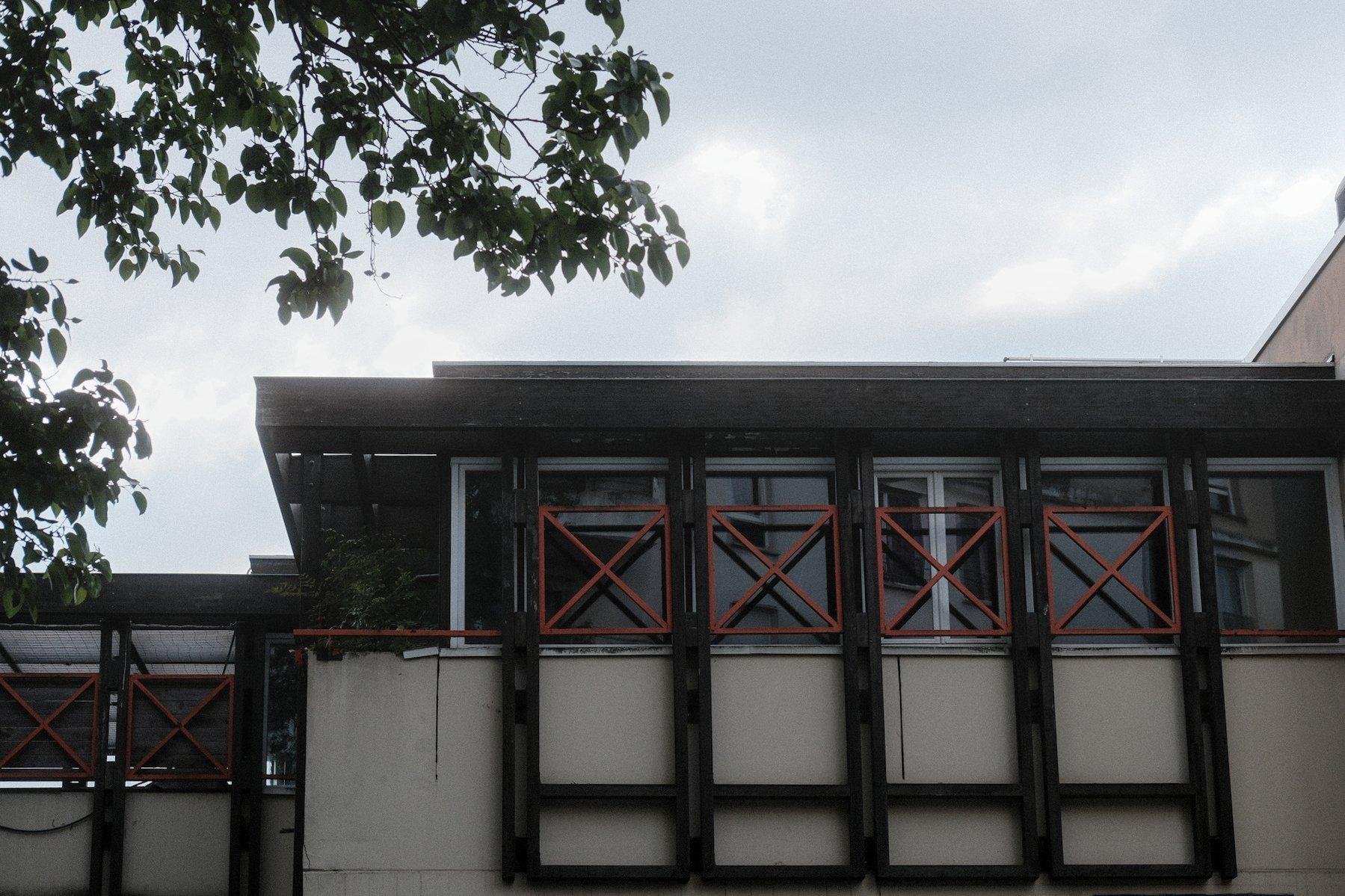 Photo horizontale. Le haut d’un bâtiment à 2 ou 3 étage au toit plat. Des fenêtres sont barrées d’une barrière rouge en forme de carré barré d’une croix