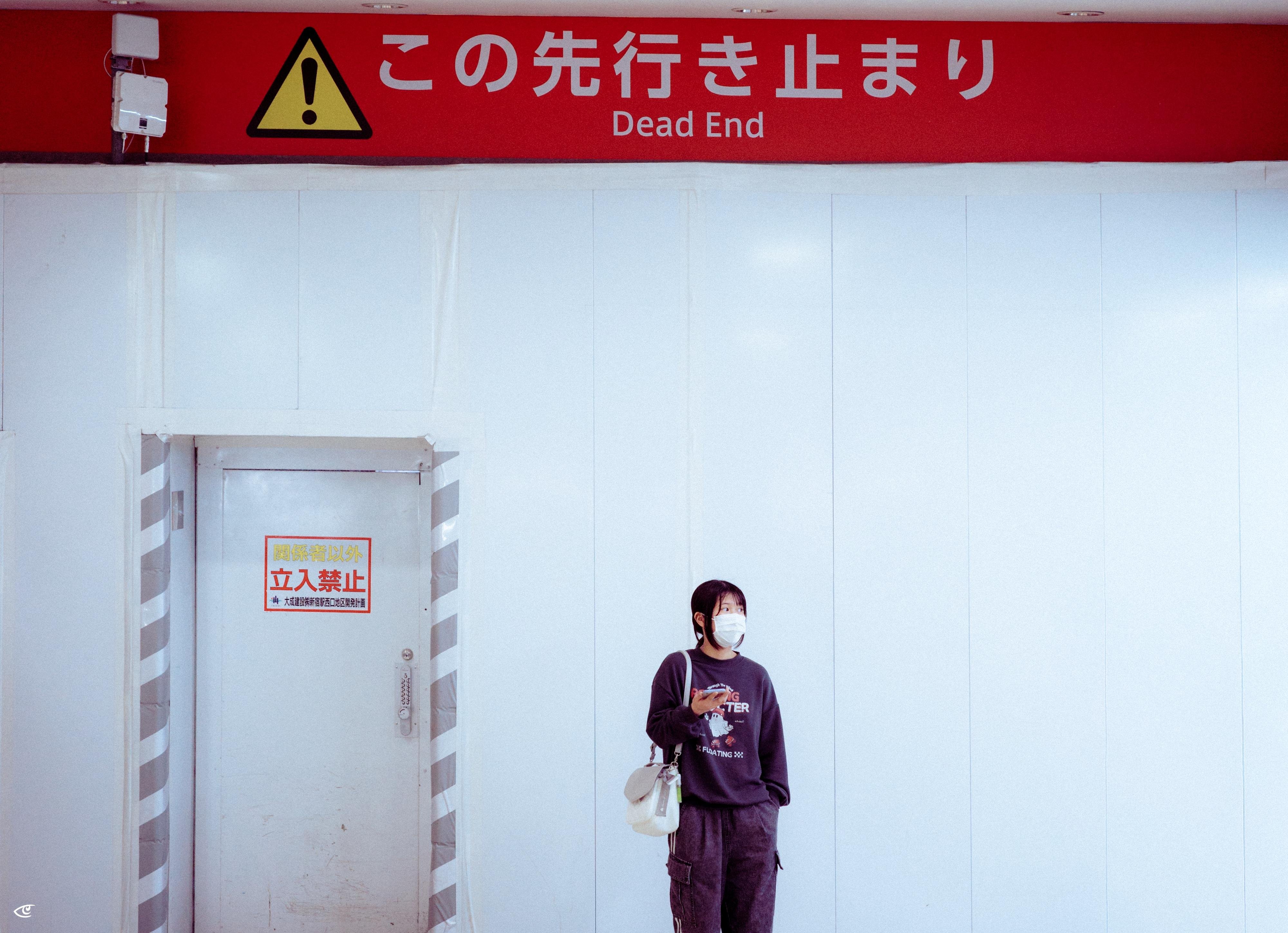 Person wearing a face mask stands against a white wall beside a closed door with a Japanese “No entry” sign; a red overhead sign with a yellow warning triangle reads “Dead End” in English and Japanese.