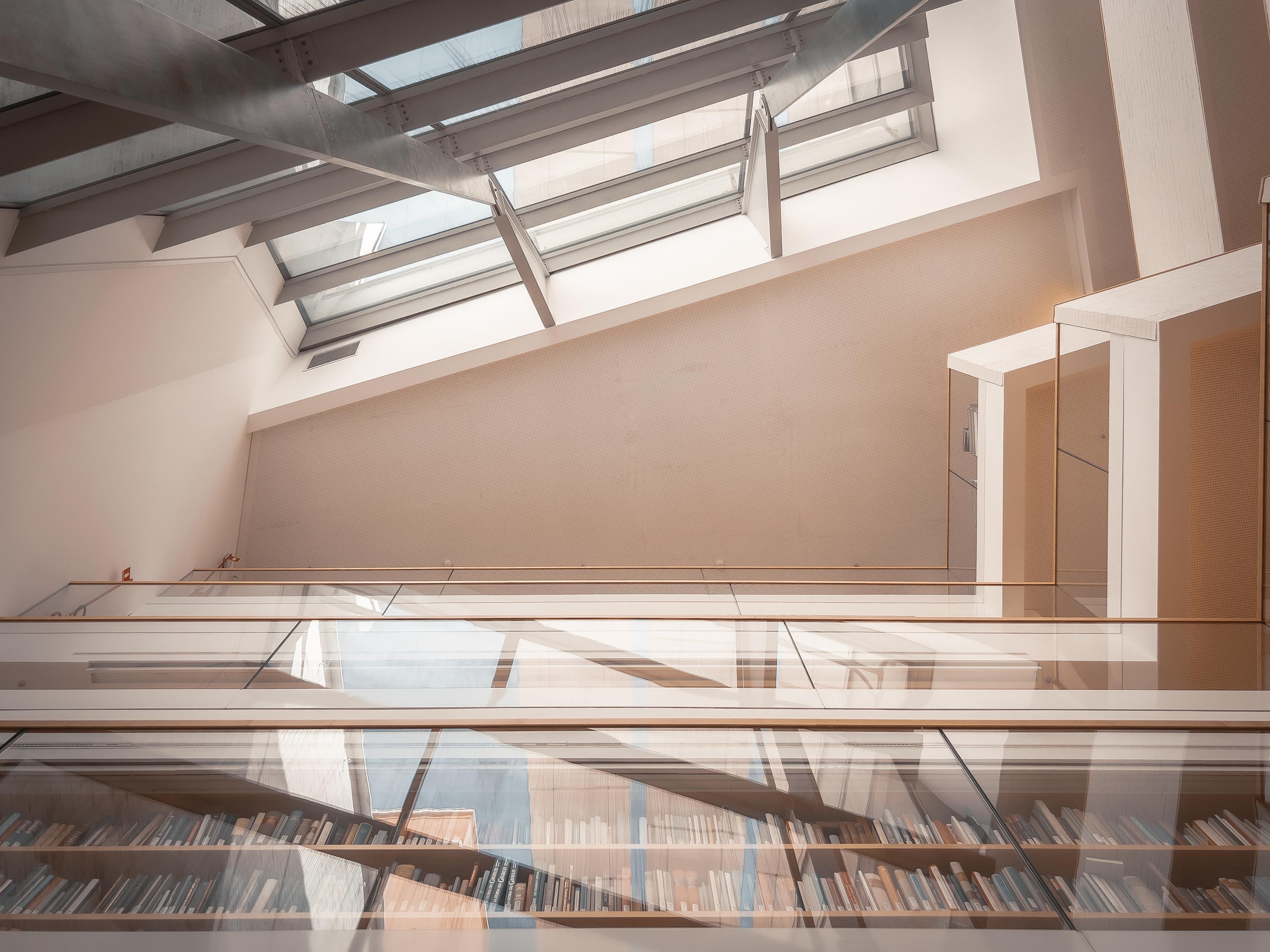 Spazio interno con un design architettonico moderno. C'è un grande soffitto in vetro o lucernario che permette alla luce naturale di inondare l'area. Sotto, c'è una serie di scaffali pieni di libri, suggerendo che potrebbe essere una biblioteca o un'area di lettura. Lo stile complessivo è pulito e contemporaneo.