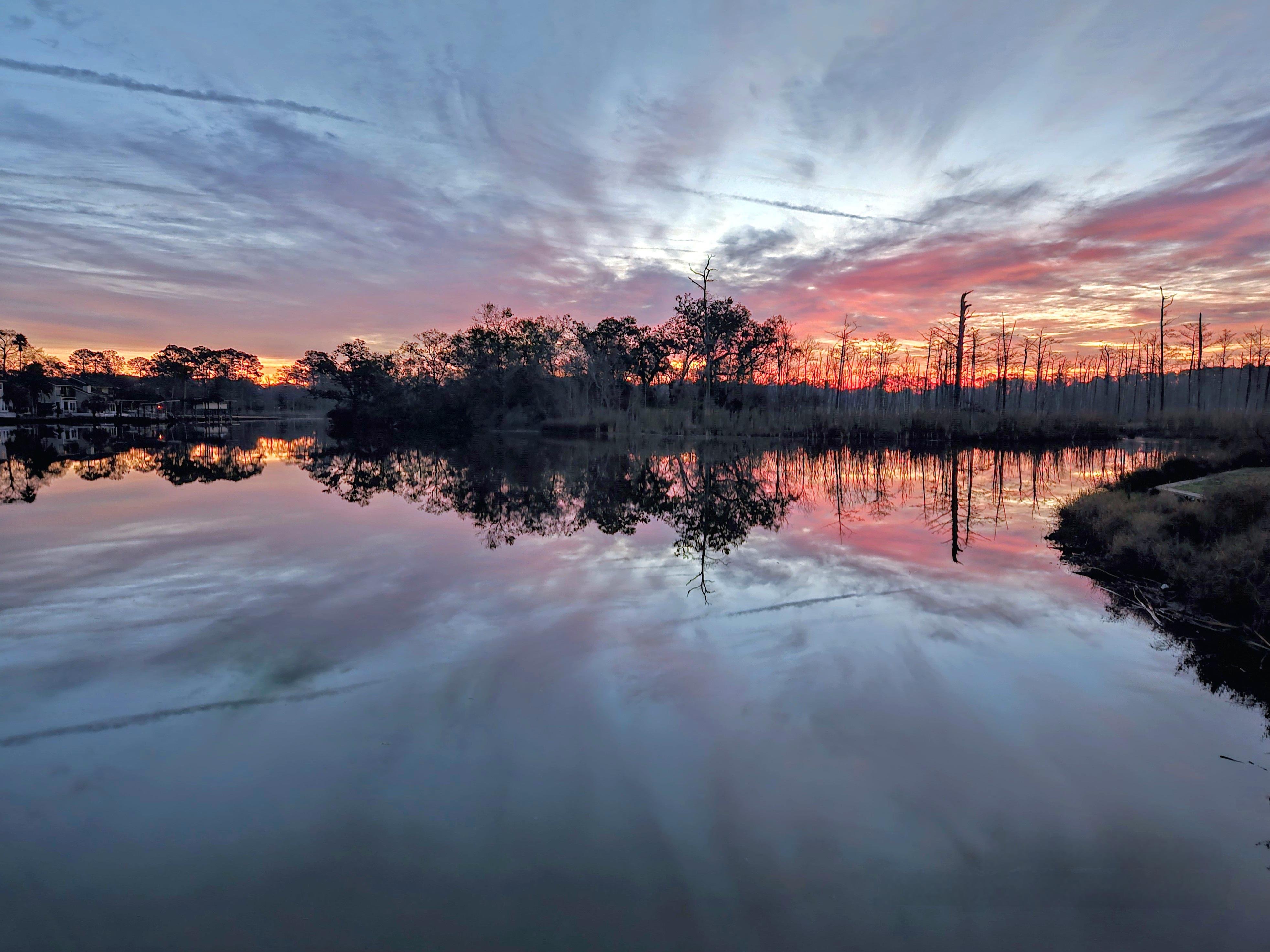 Sunrise over a calm waterway with a sky filled with purple, pink, and orange hues. Trees line the horizon, their silhouettes reflected in the water, creating a symmetrical effect. Thin wispy clouds spread across the sky, with a darker foreground on the right displaying patches of grass.