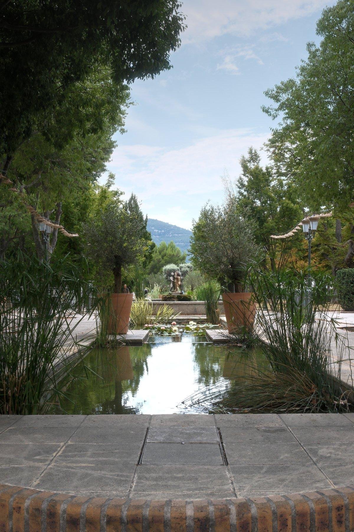 An artificial pond, probably a fountain, surrounded by trees and plants. Midway, 2 giants flower pots with small trees in them, at the far end, a statue and in the background, mountains, under a blue sky with some white clouds