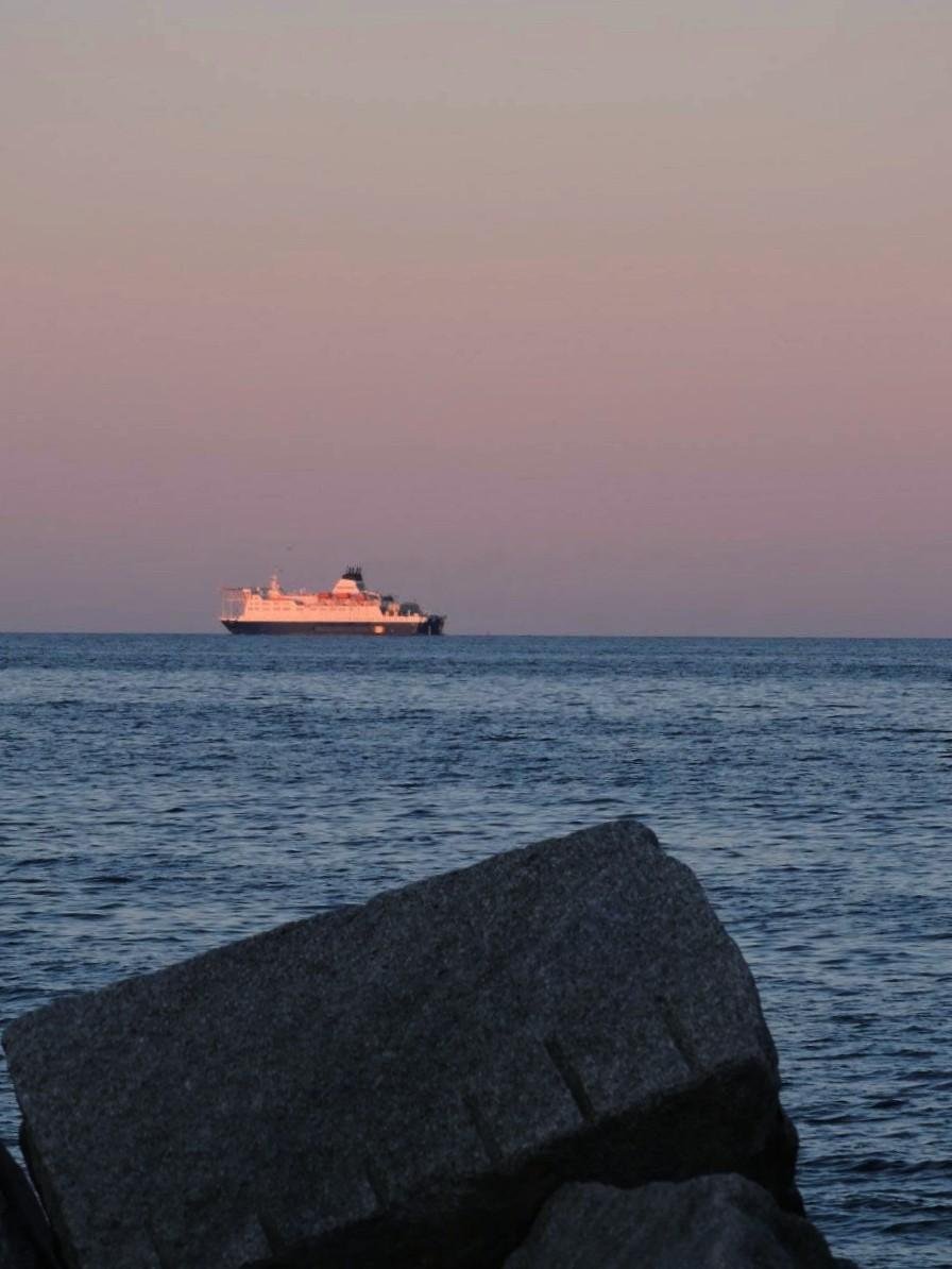 Ferry sailing across a calm sea under a pinkish sky at sunset, with large rocks in the foreground.