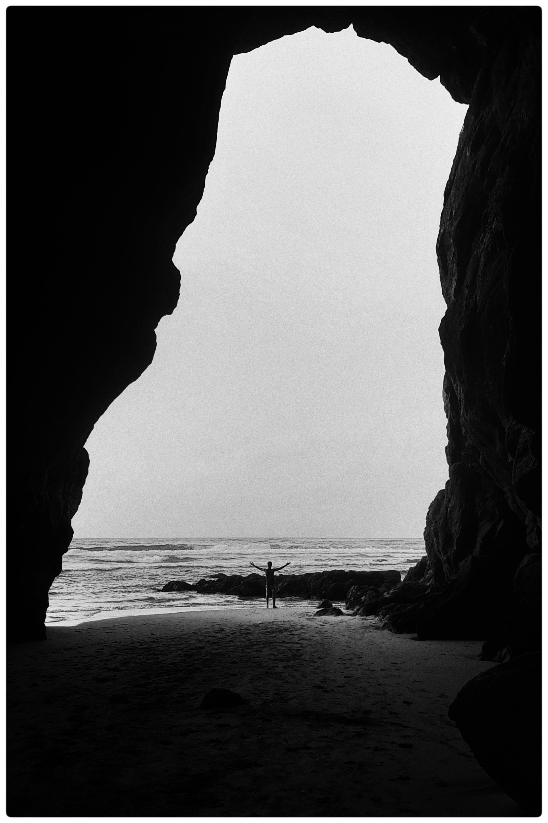 Silhouette of a person standing with arms outstretched on a sandy beach, framed by a large dark sea cave opening, with rocks and ocean waves beyond under a bright sky.