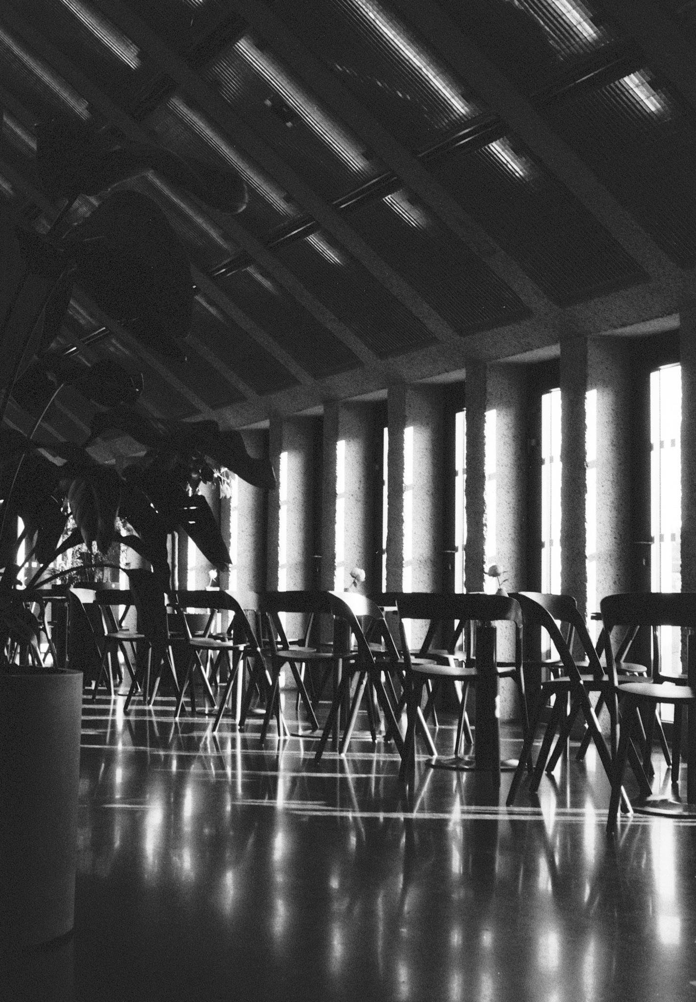 Black-and-white interior of a large hall with a slanted metal-beam ceiling, tall rectangular windows along the right wall, rows of empty folding chairs, and a potted plant in the left foreground; light from the windows reflects on the glossy floor.