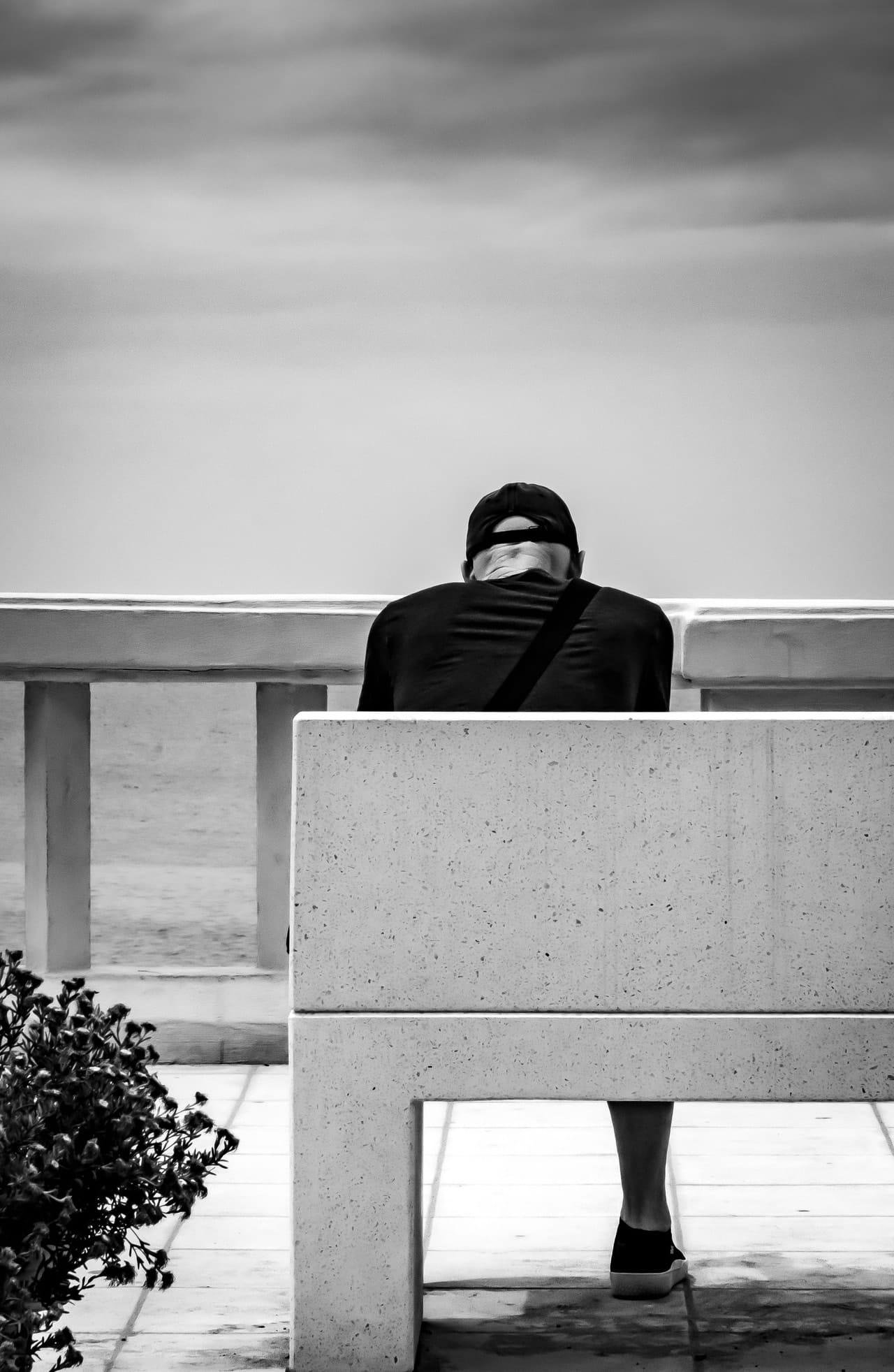 Black-and-white photo of a person wearing a backward cap and dark shirt, seated on a stone bench and leaning forward near a seaside railing; ocean and cloudy sky in the background, with a small shrub in the lower left.