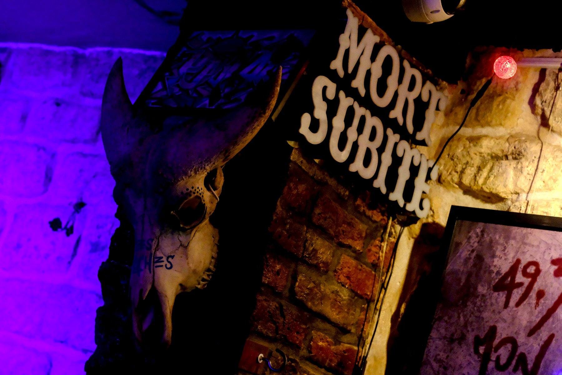 Photo horizontale. Sur la tranche d’un petit bout de mur perpendiculaire au mur principal, un crâne de taureau est accroché. Sur la face qu’on voit de ce bout de mur, une décoration composées de lettre à la typo évoquant le Far West écrit « Mort Subite »