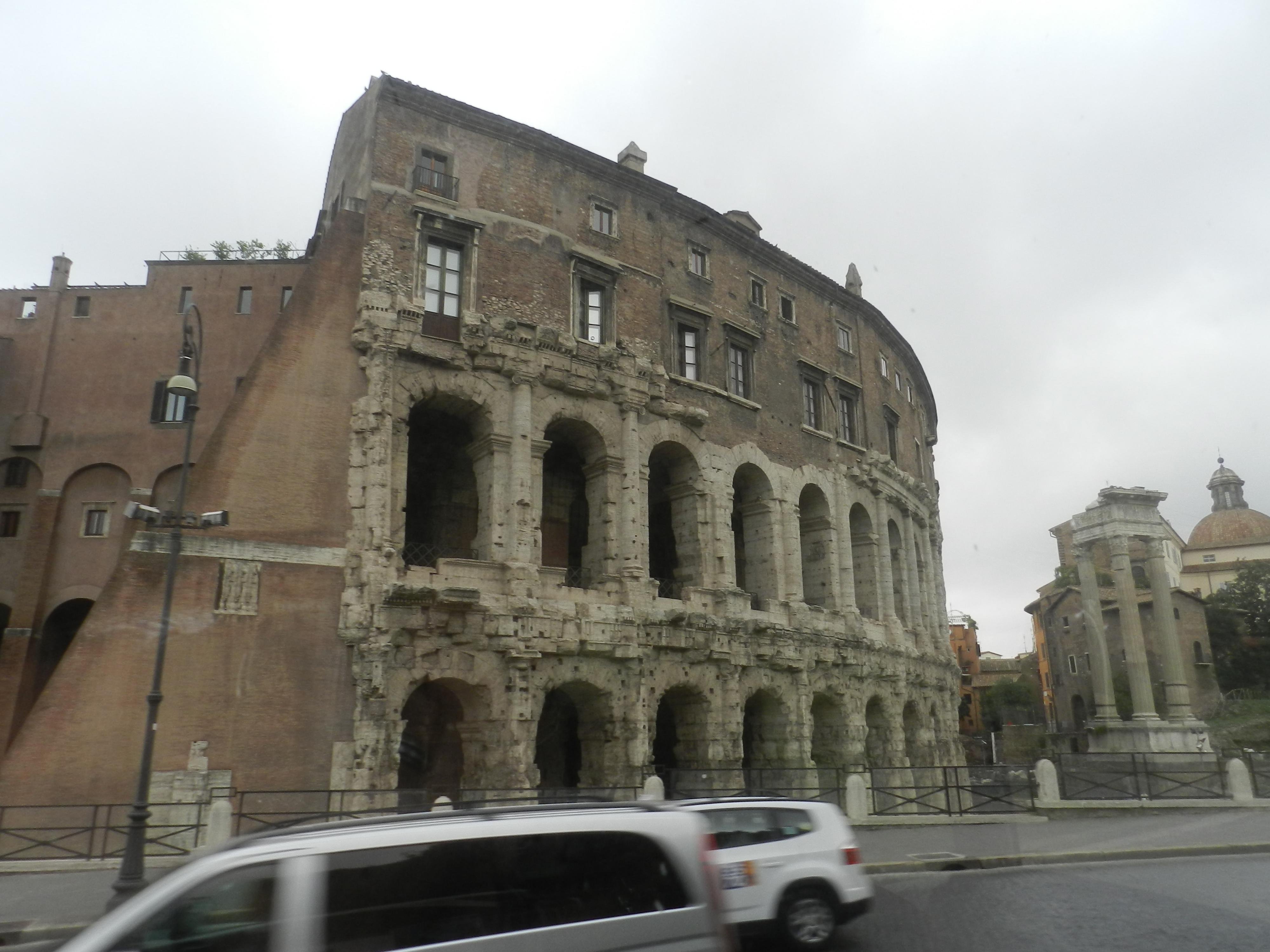 A photo taken from a moving tour bus in Rome shows the massive, weathered stone façade of the Teatro di Marcello (Theatre of Marcellus), an ancient open-air theatre commissioned by Julius Caesar and completed under Augustus around 13 BCE. The structure features two preserved tiers of large arches built from travertine, with later medieval and Renaissance buildings rising directly above them, giving the theatre its distinctive layered look. The cloudy sky casts a soft grey light over the scene. In the foreground, cars blur past, emphasizing the movement of the bus. To the right stands the Temple of Apollo Sosianus, identifiable by its three standing Corinthian columns that remain from the original Republican temple. Behind it, an ochre-colored church dome peeks above the rooftops. The image captures a fleeting yet striking moment where ancient Roman architecture and modern city life coexist side by side as the tour heads toward its starting point.