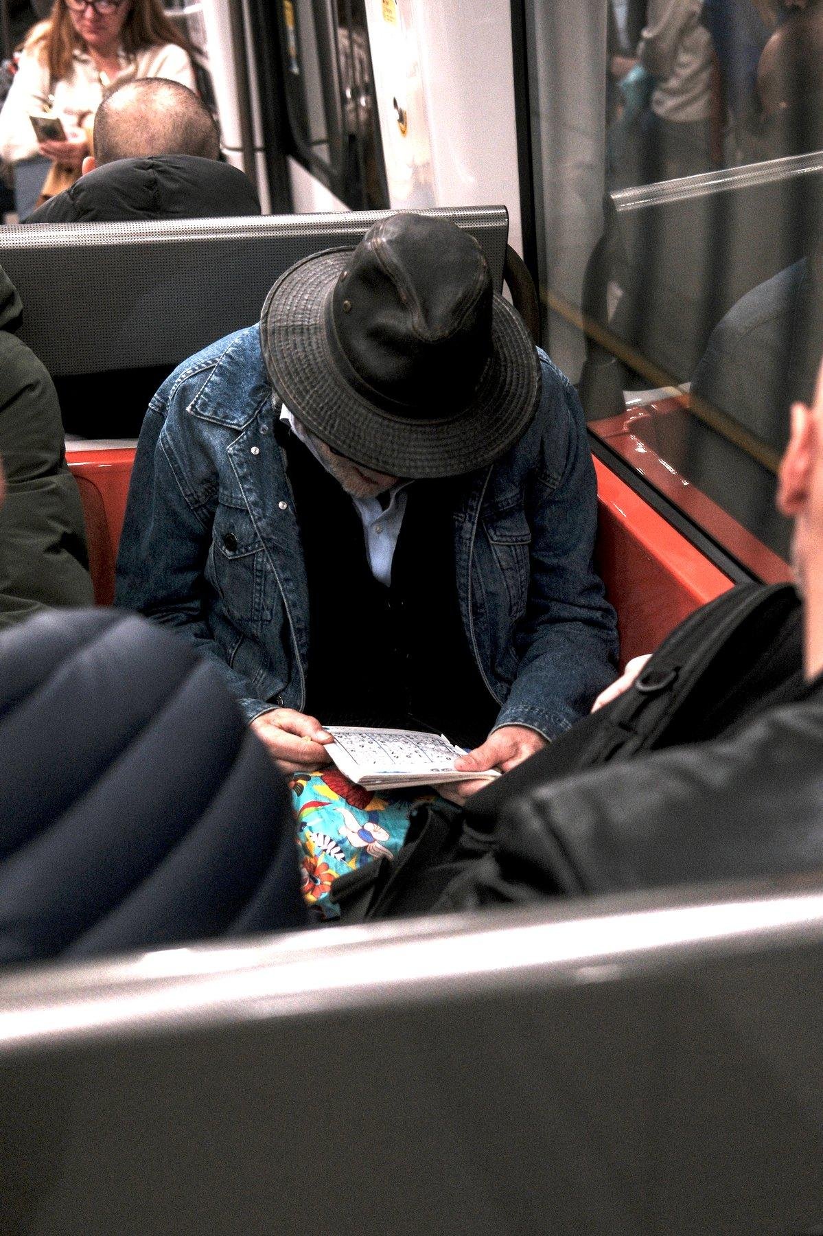 Photo verticale. Un homme dont le visage est caché par un chapeau en cuir noir, portant une chemise bleu clair, un gilet noir et une veste en denim. Il est assis dans le métro et a la tête penchée sur un carnet de Sudoku