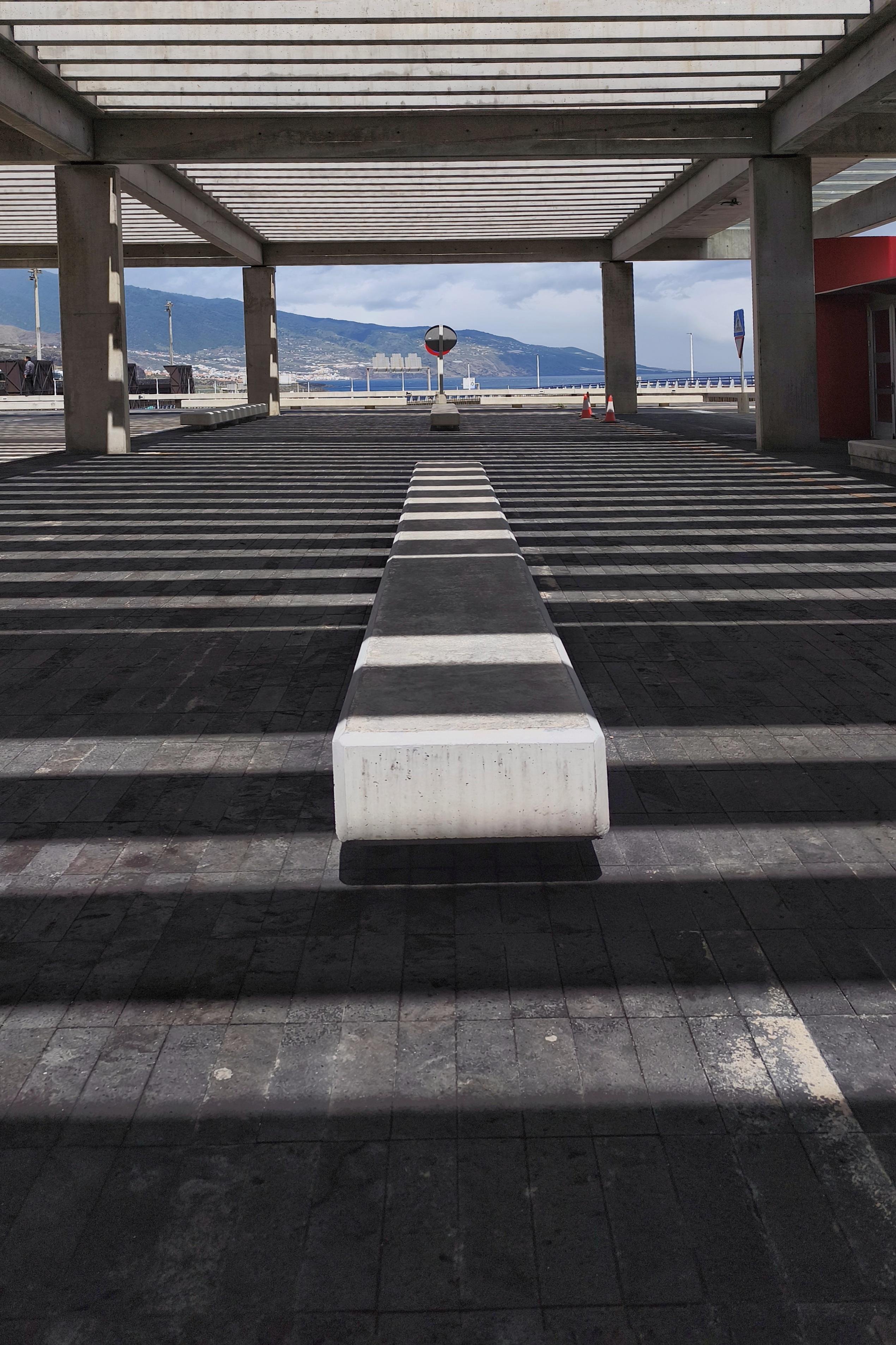 Covered parking area with concrete beams casting striped shadows on the pavement, a long rectangular concrete barrier centered in the foreground, and a circular road sign and orange cones near the open edge overlooking water and distant mountains. 
### 
Überdachter Parkplatz mit Betonbalken, die streifenförmige Schatten auf den Asphalt werfen, eine lange rechteckige Betonbarriere in der Mitte im Vordergrund und ein rundes Verkehrsschild und orangefarbene Kegel in der Nähe des offenen Randes mit Blick auf Wasser und ferne Berge.