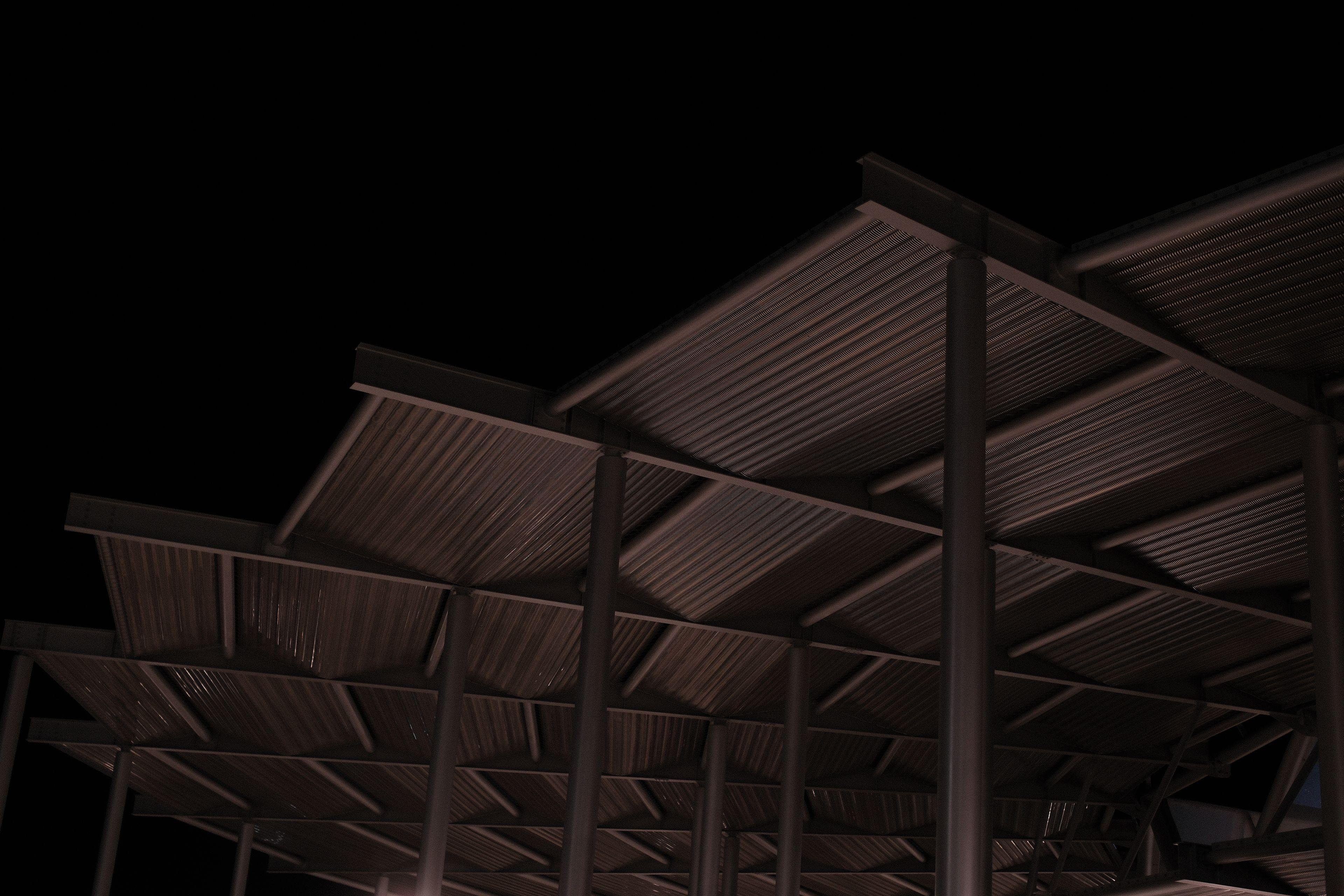 Photo format paysage, de nuit, montrant la toiture en métal, non régulière d’un préau formant un bout de spirale, éclairé par en dessous d’une lumière orangée, sur fond de ciel noir