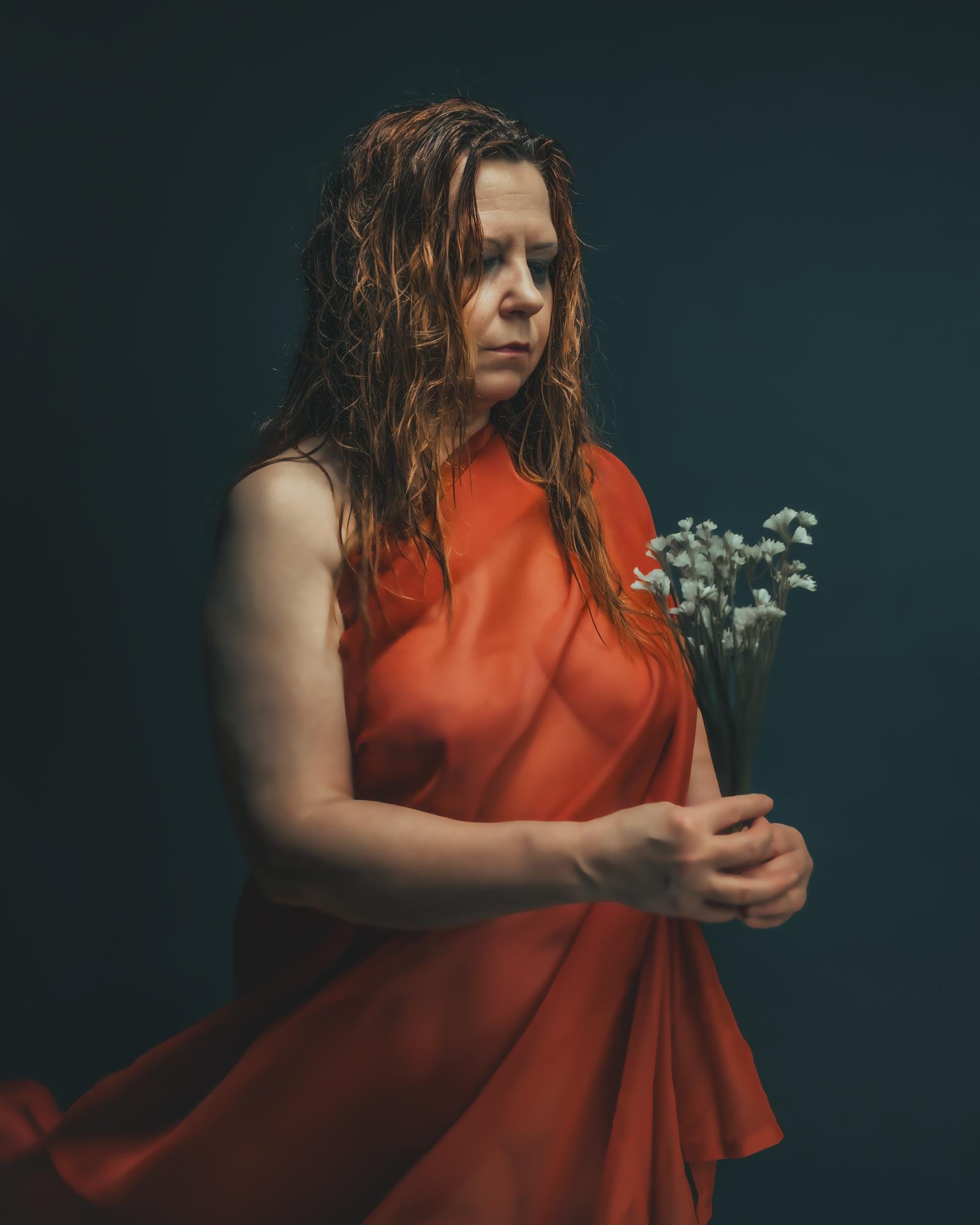 The image shows a person wearing a red draped garment, holding a small bouquet of flowers. The background is a dark, solid color, and the person appears to have wet hair and is gazing downward, creating a serene or contemplative mood.