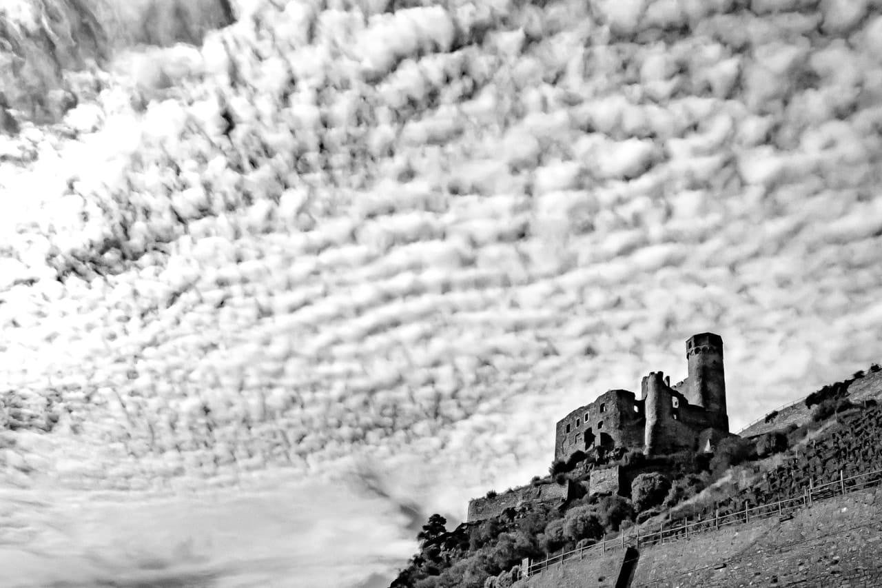 Black-and-white landscape with a ruined stone castle and tall round tower on a hillside at right, beneath a sky filled with dense rippled cloud bands; sloping terrain with vegetation and a fence in the foreground.