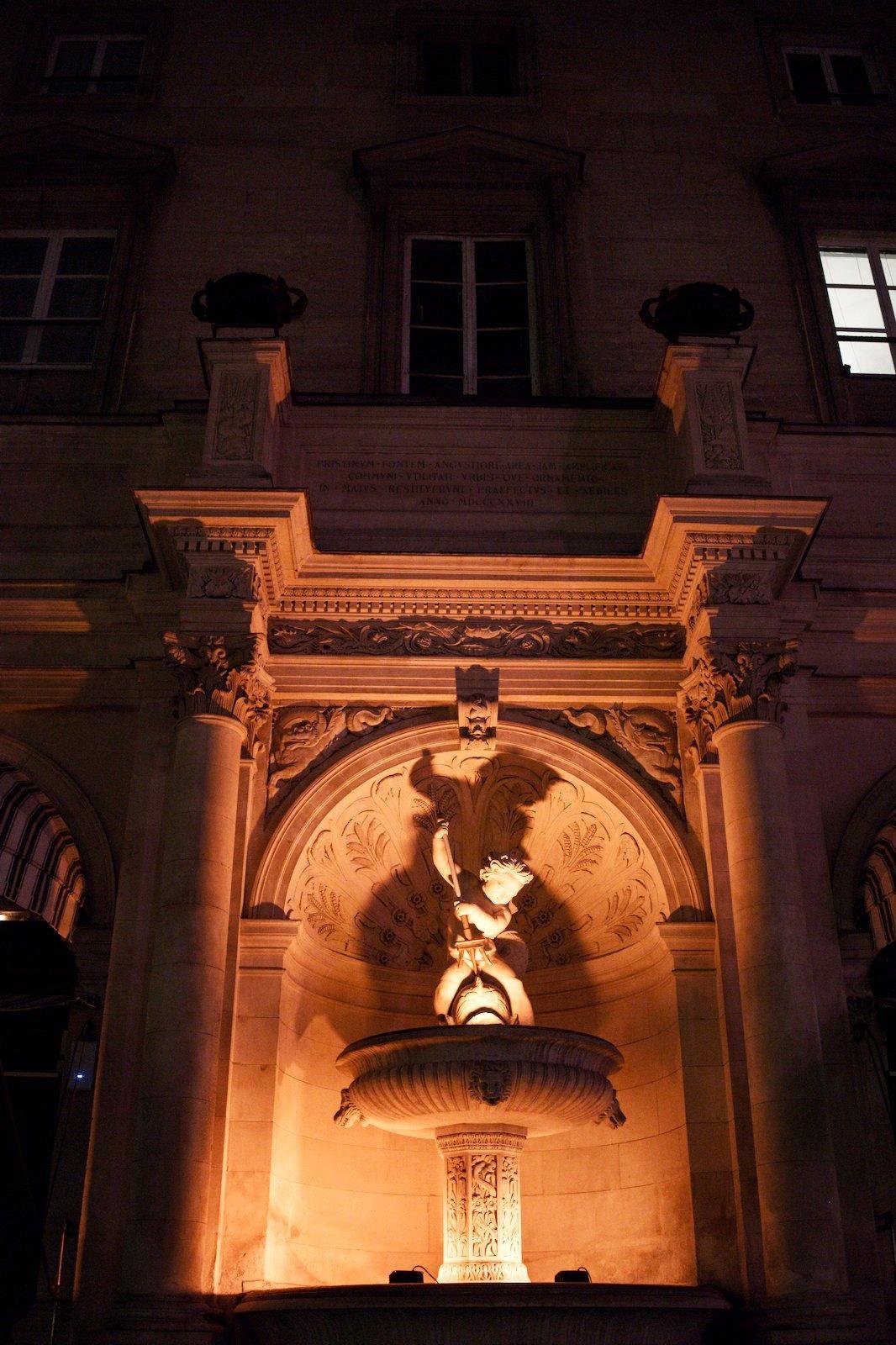 Photo verticale. Une alcove dans une façade d’immeuble, contenant une fausse (?) fontaine en pierre surplombée d’une statue d’angelot qui tiens un trident et s’apprête à l’enfoncer dans un poisson. Le tout est éclairé par le bas, de nuit, projetant l’ombre de l’angelot sur le plafond de l’alcôve.