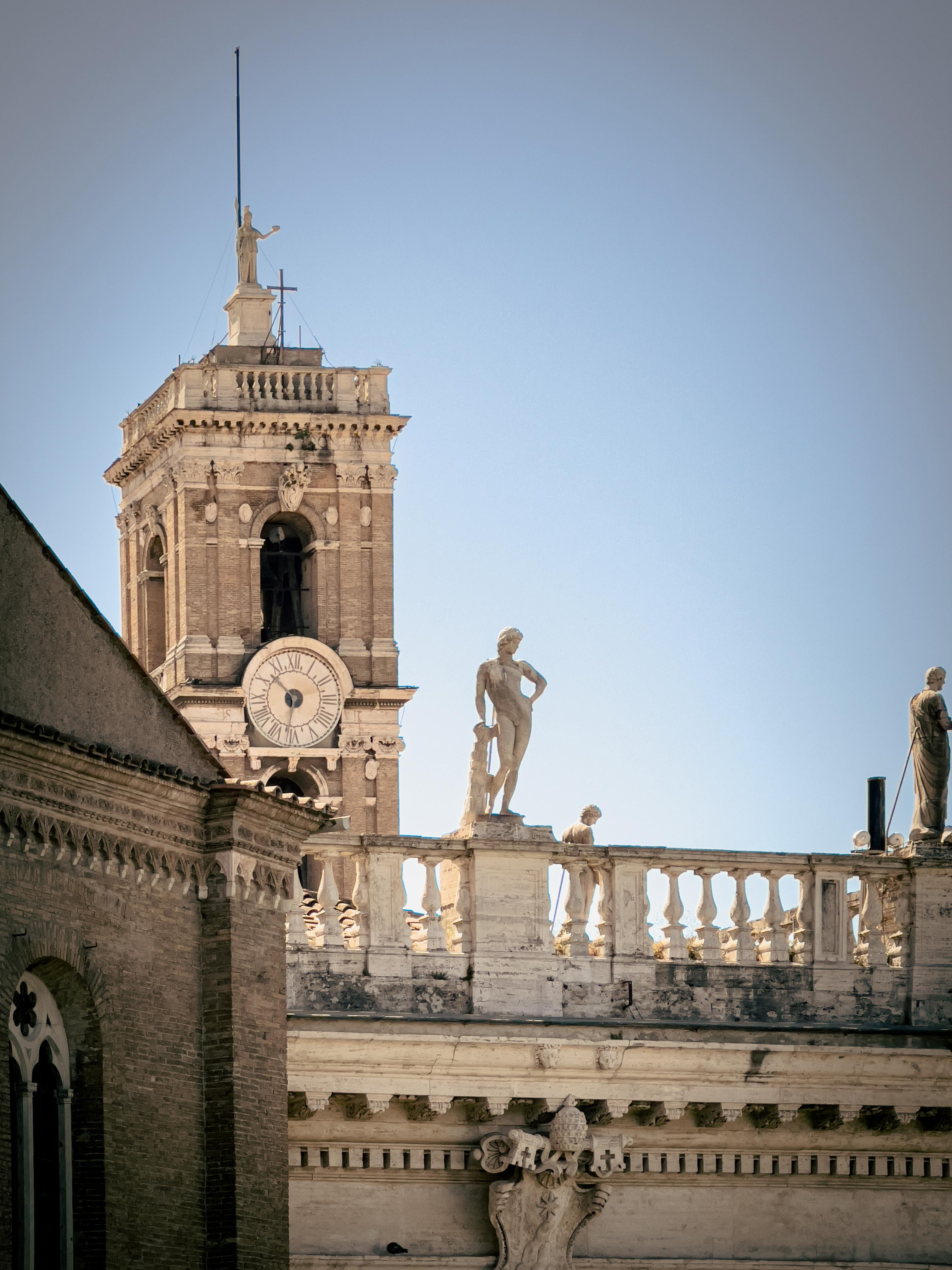 Veduta architettonica con una torre dell'orologio e statue su una balaustra. L'edificio presenta elementi classici, come colonne e intagli ornati. La torre dell'orologio è sormontata da una statua e lo stile complessivo suggerisce un'influenza architettonica storica o europea.