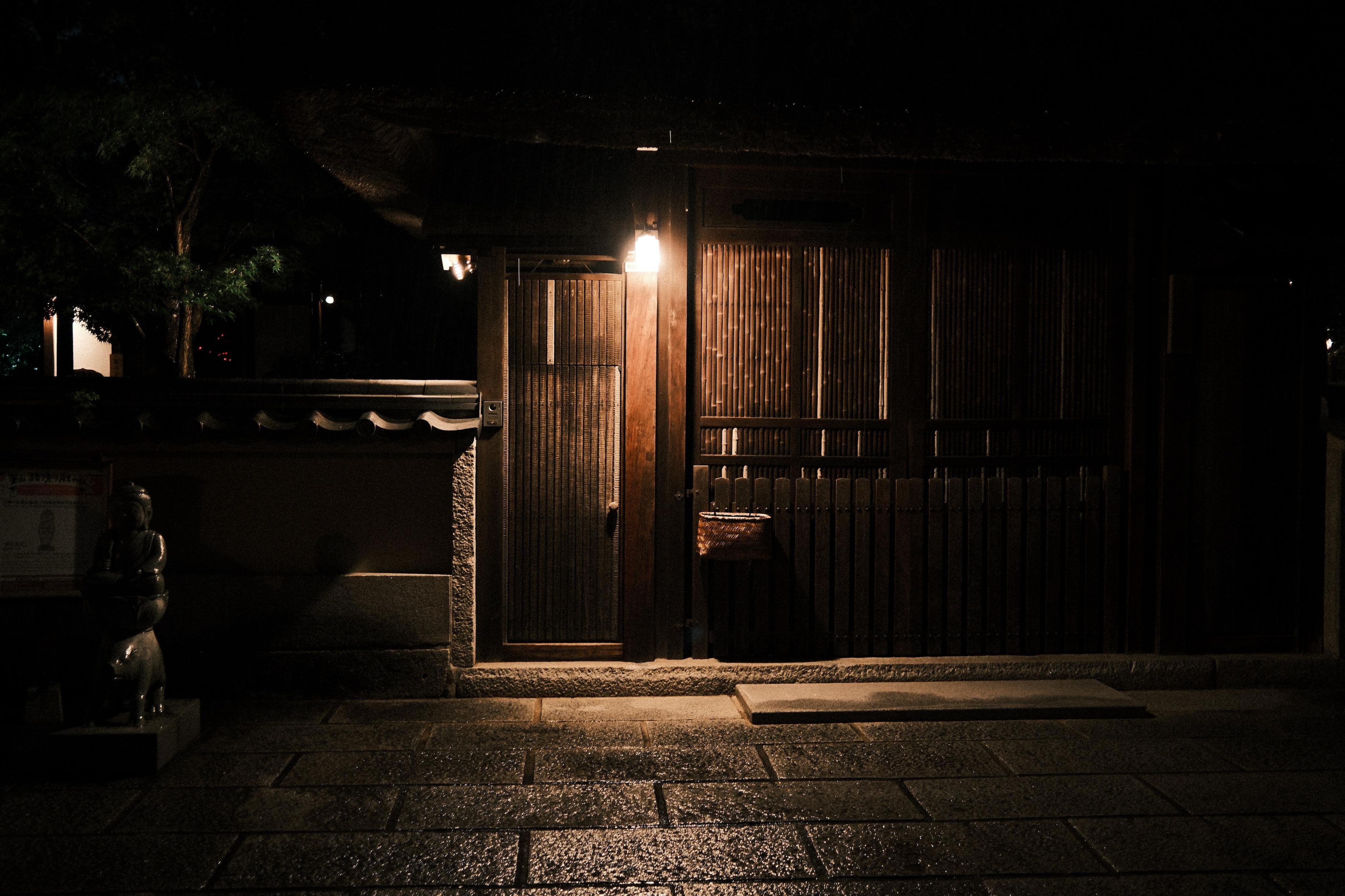 Dimly lit wooden gate and slatted wall of a traditional building at night, illuminated by a single wall lamp; wet stone-paved walkway in the foreground with a small statue and low wall on the left.