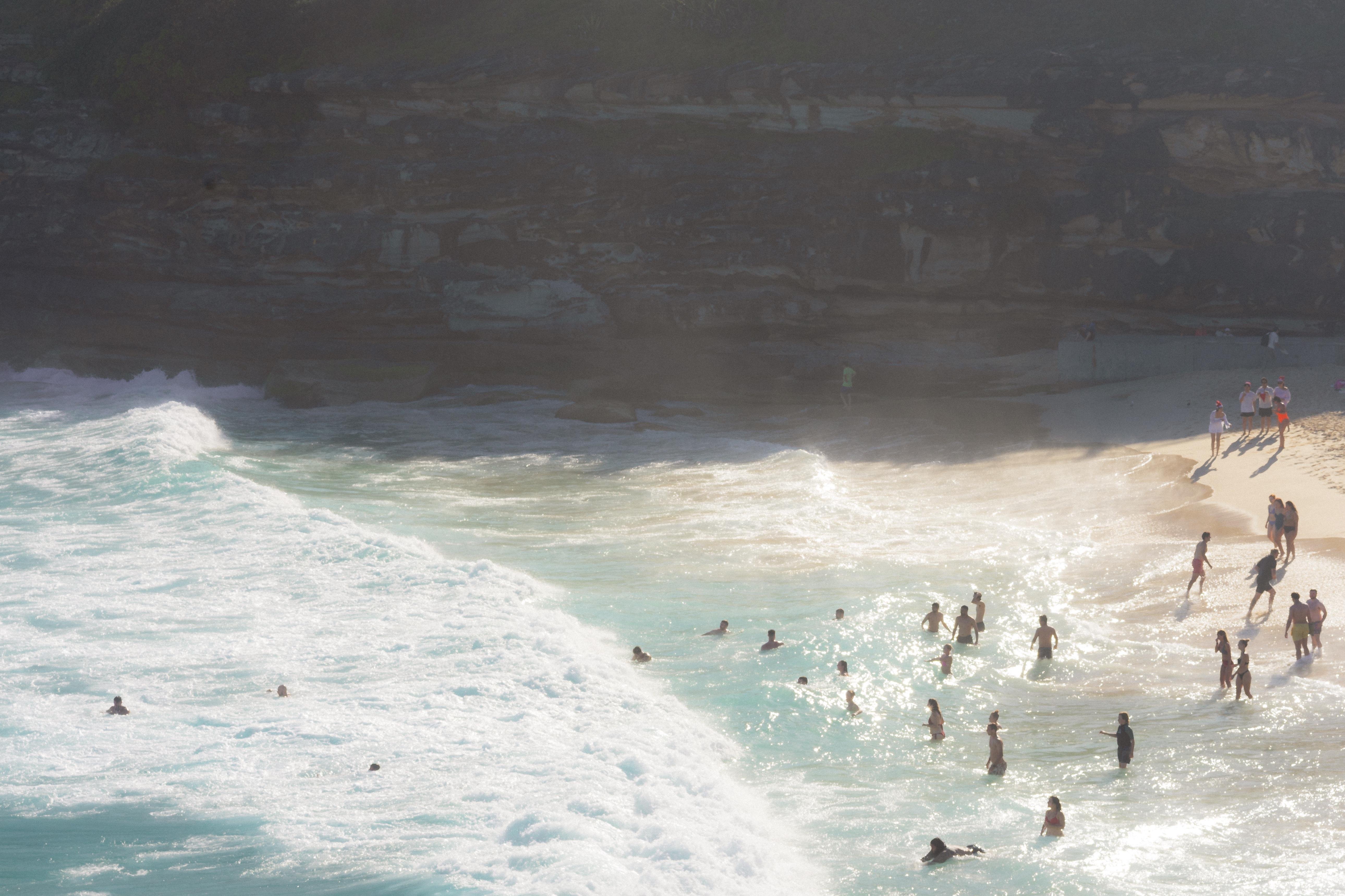 The image depicts a beach scene with people swimming and playing in the ocean. The water is a bright blue, with waves crashing near the shore. On the sand, several groups of people are walking or standing. The image appears to have a warm, sunny atmosphere.