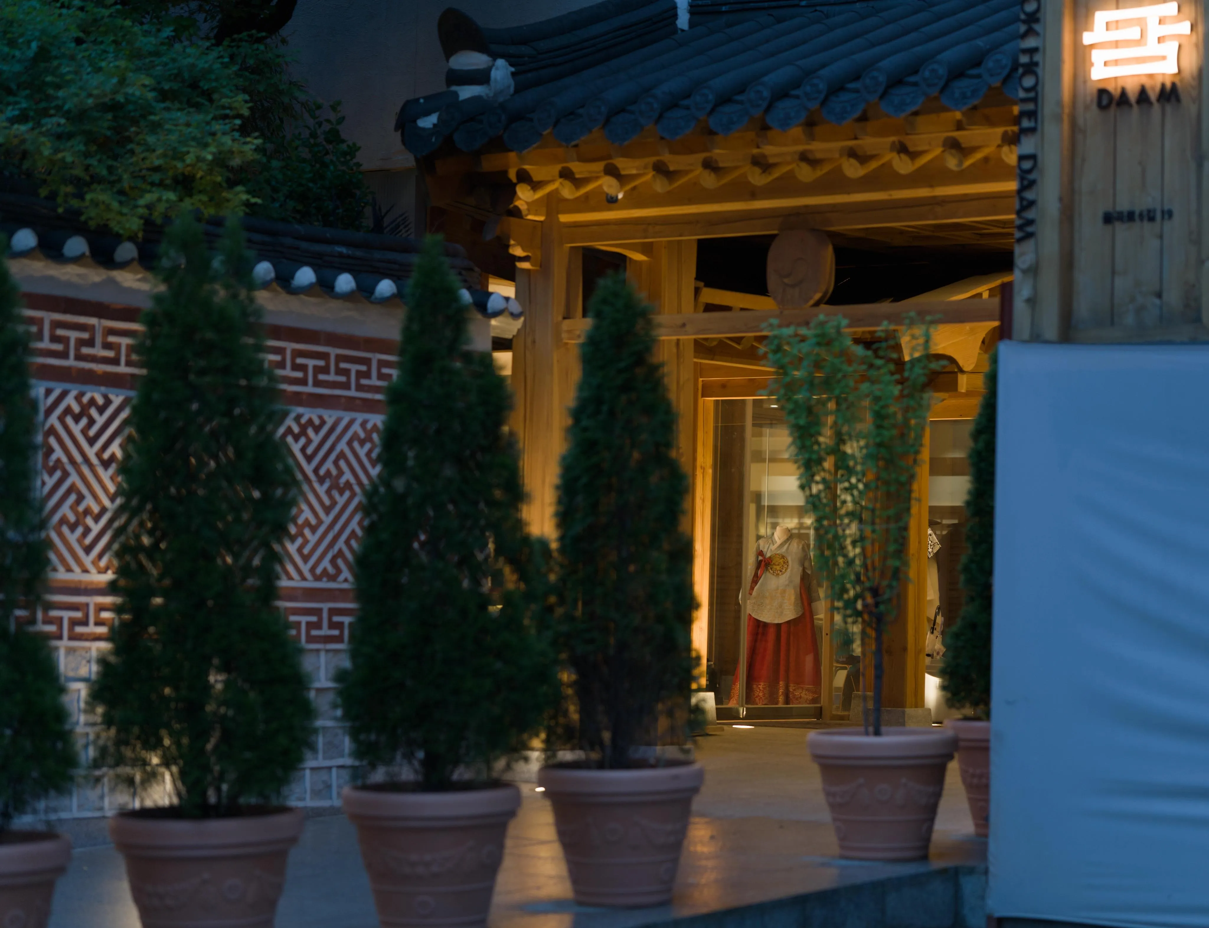 Photograph of the entrance of a building built as wooden gate in a traditional Korean architectural style, with a traditional white and red woman's dress on a mannequin inside the front window. The depth-of-field focuses on the mannequin. The wooden gate entrance is lighted in yellow, while the exterior has the blue tones of night photography. A sign indicates that the building is a hotel. Around the wooden gate-style entranceway is a traditional-looking patterned wall. In the foreground out of focus, small potted trees are lined up in front of the hotel entrance.