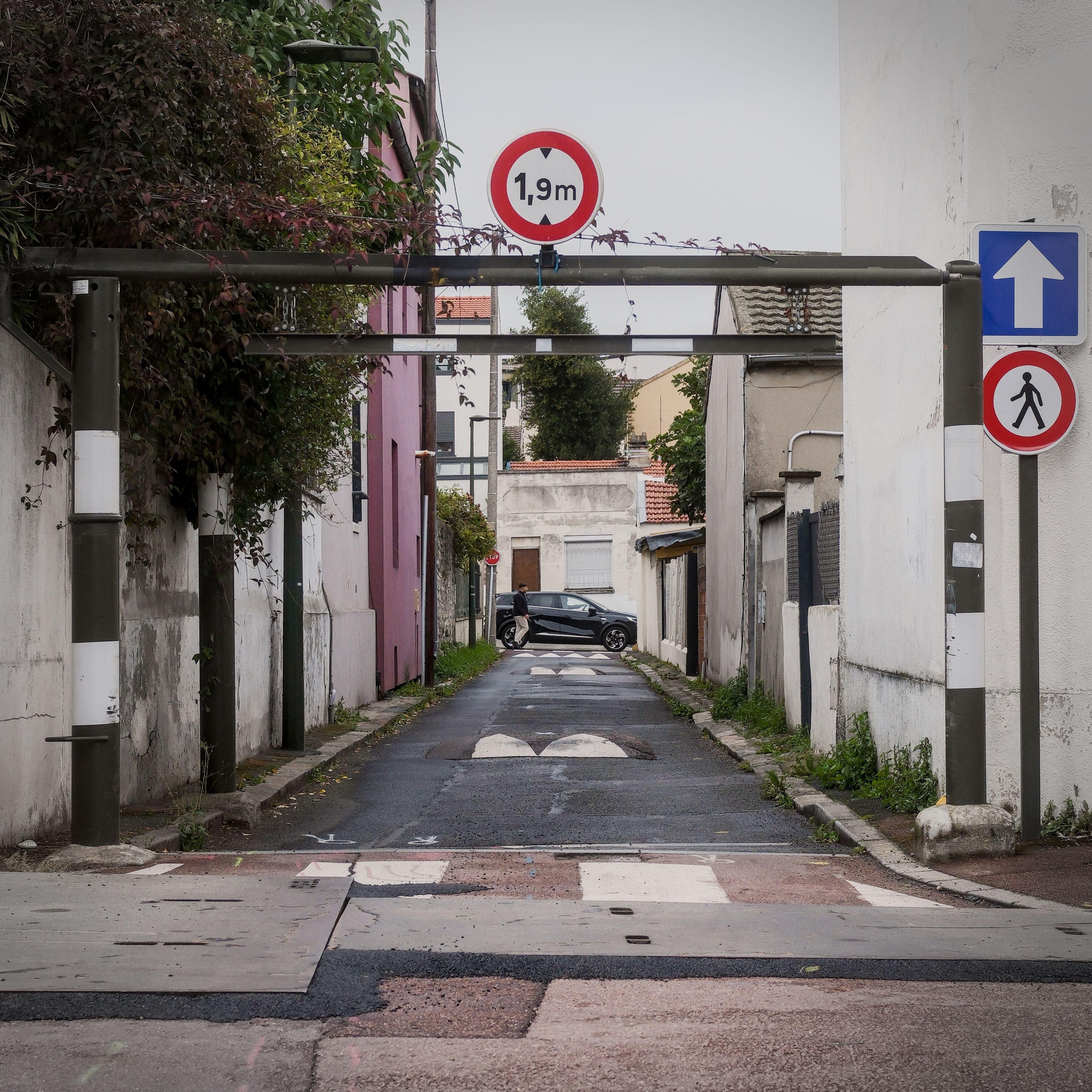 Photo carrée. Une petite rue dont l’entrée possède un cadre en métal avec un panneau hauteur limitée à 1,9m au dessus. La rue a aussi des ralentisseurs et est assez étroite, juste une voiture peut passer et ce qui fait office de trottoir est juste assez large pour un lampadaire.