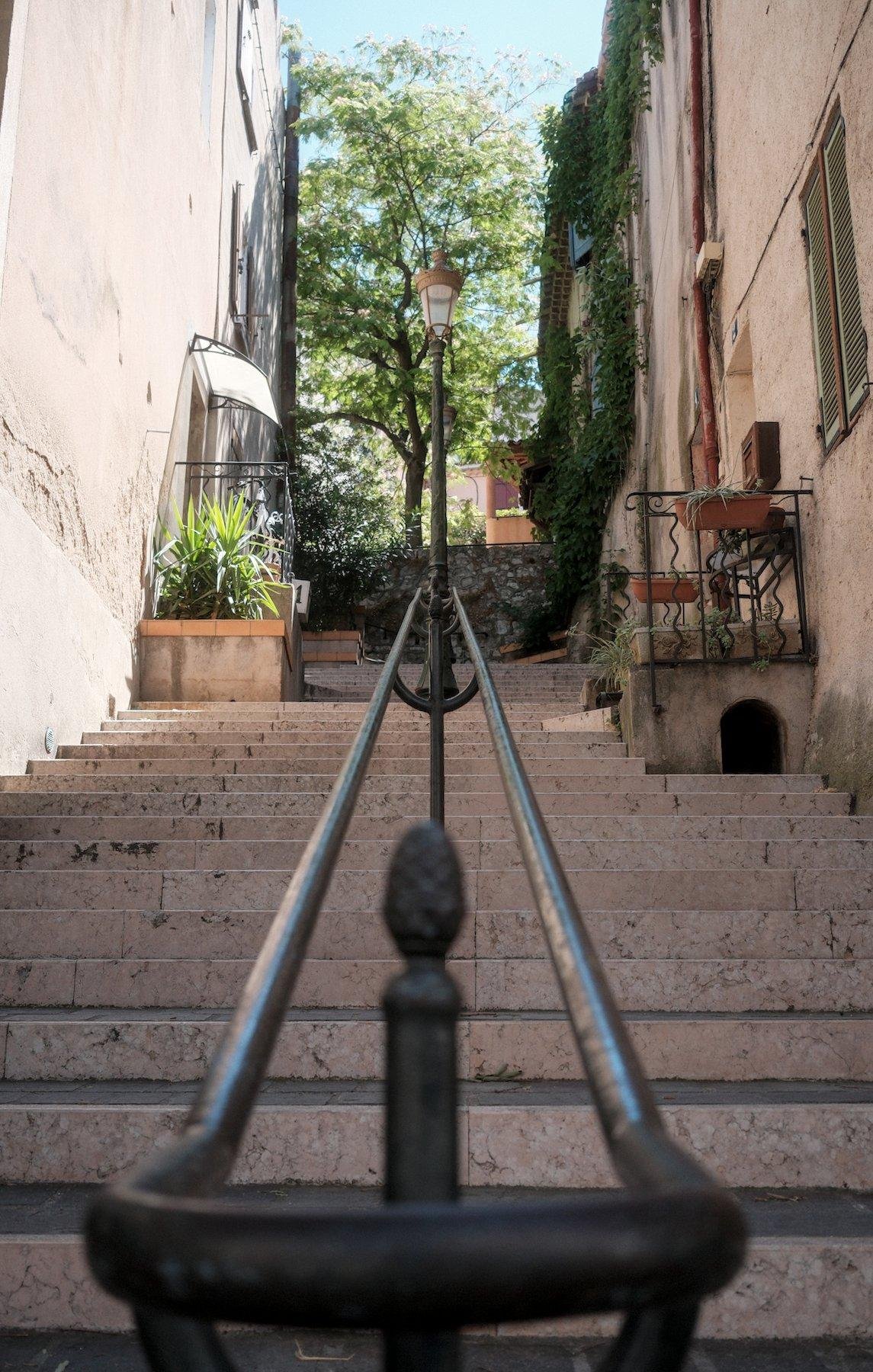 Photo verticale. Vue à ras d’une double rampe d’escalier centrale, à l’extrémité basse où la rampe fait un arc de cercle pour aller de l’autre côté. On voit Les escaliers montant entre 2 rangés de maisons, un grand arbre tout au bout