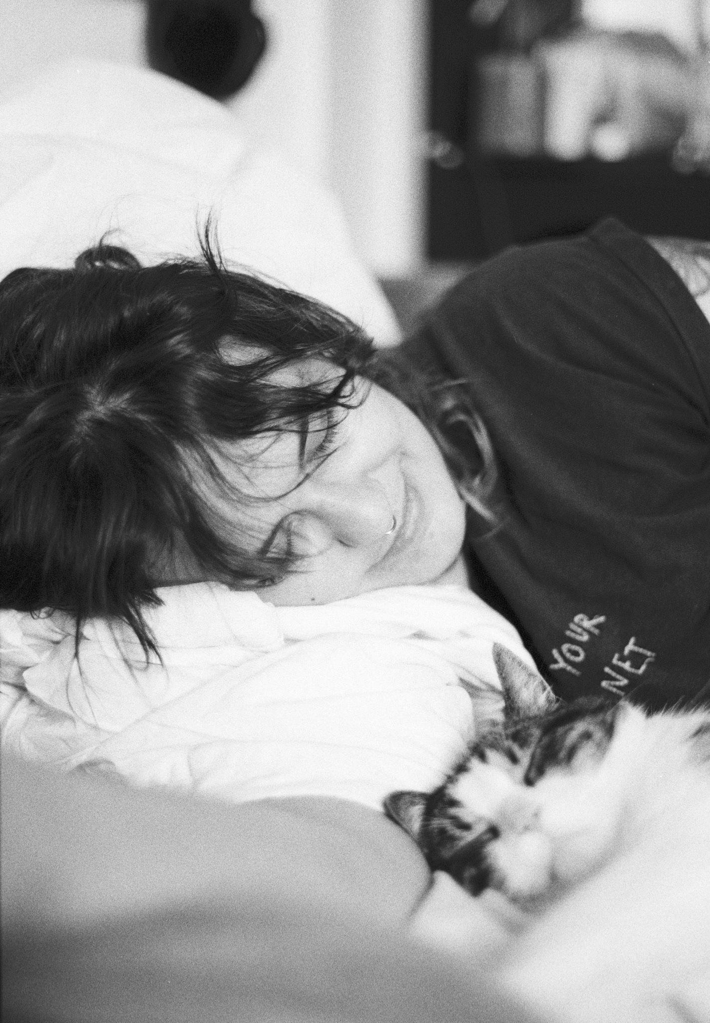 Person with tousled dark hair lying on a bed with their head on a pillow, eyes closed, wearing a dark T‑shirt with partial text “YOU’R…NET,” next to a tabby-and-white cat curled up in the foreground; black-and-white photo with a blurred background.