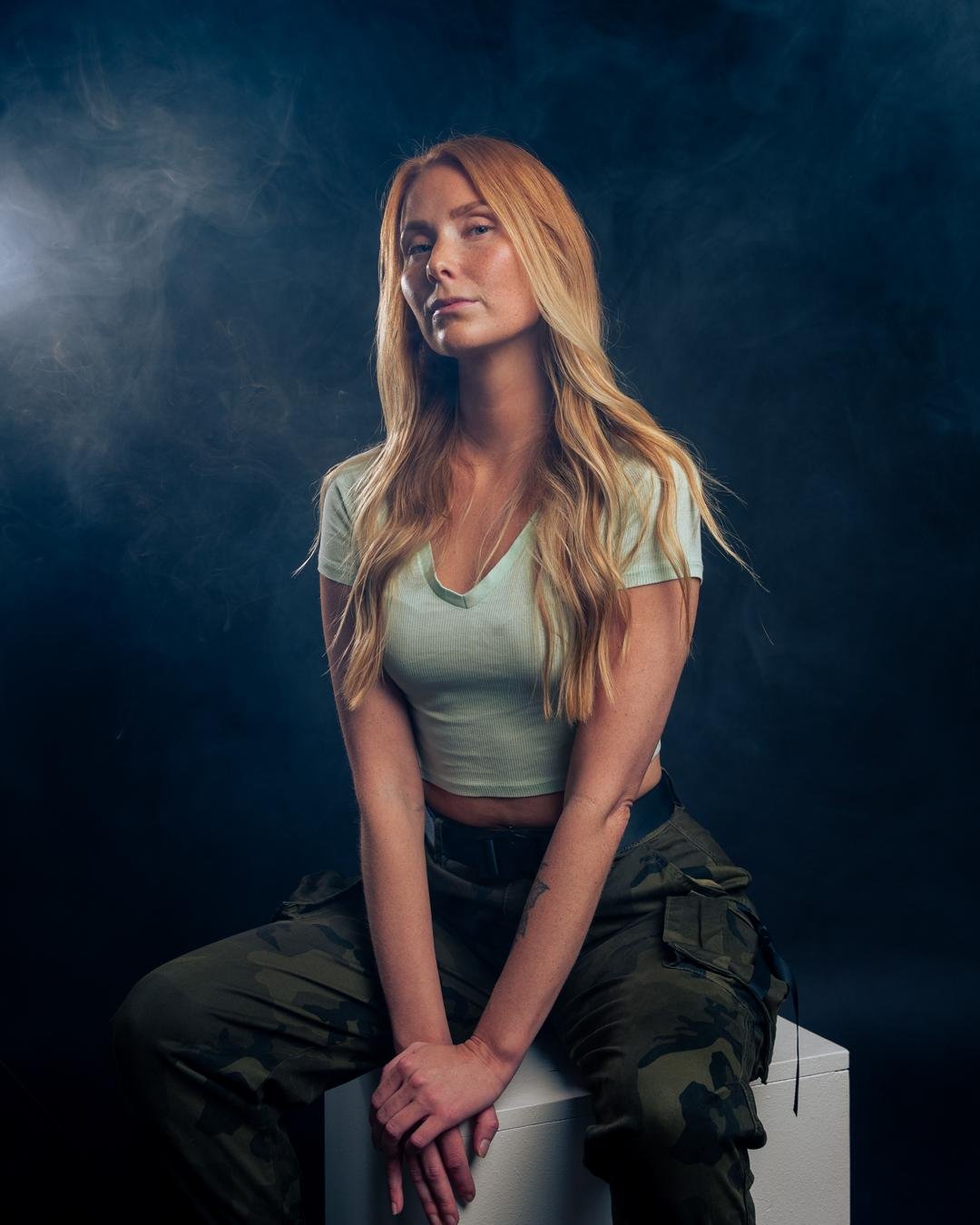 The image shows a person with long hair sitting on a white block in a dimly lit setting. They are wearing a light green top and camouflage pants. There's a moody atmosphere with a smoky or foggy background.