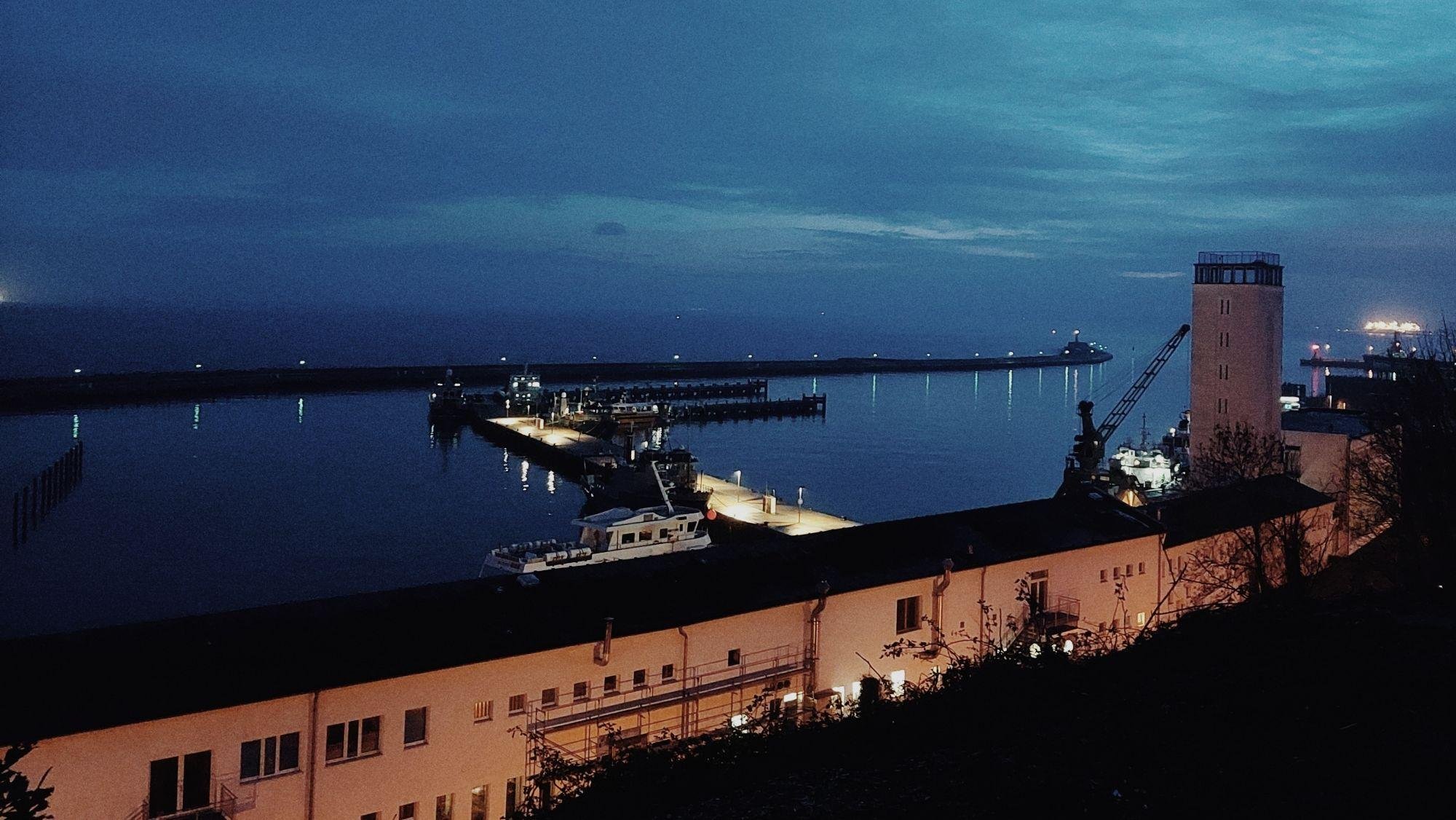 The image shows a nighttime view of a harbor or port area. There are several docks extending into the water, with lights lining the piers and illuminating the scene. Some boats or ships are docked, and buildings are visible in the foreground. The sky is dark with a hint of blue, suggesting either dawn or dusk lighting.
