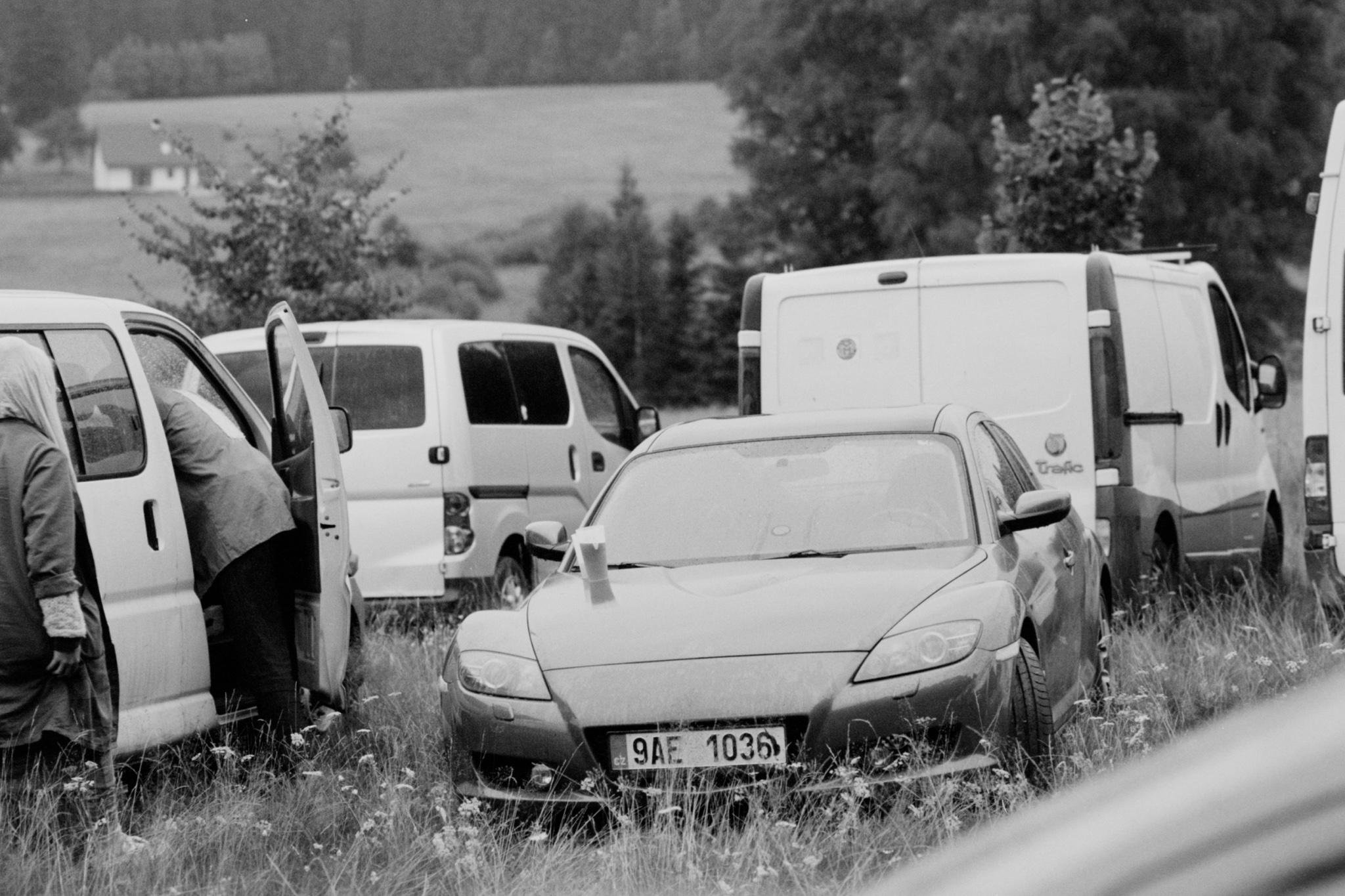 Black-and-white photo of a car partially sunk in tall grass with a small cone on its hood, surrounded by several parked vans on a rural roadside; two people stand near an open van door on the left, with trees and fields in the background.