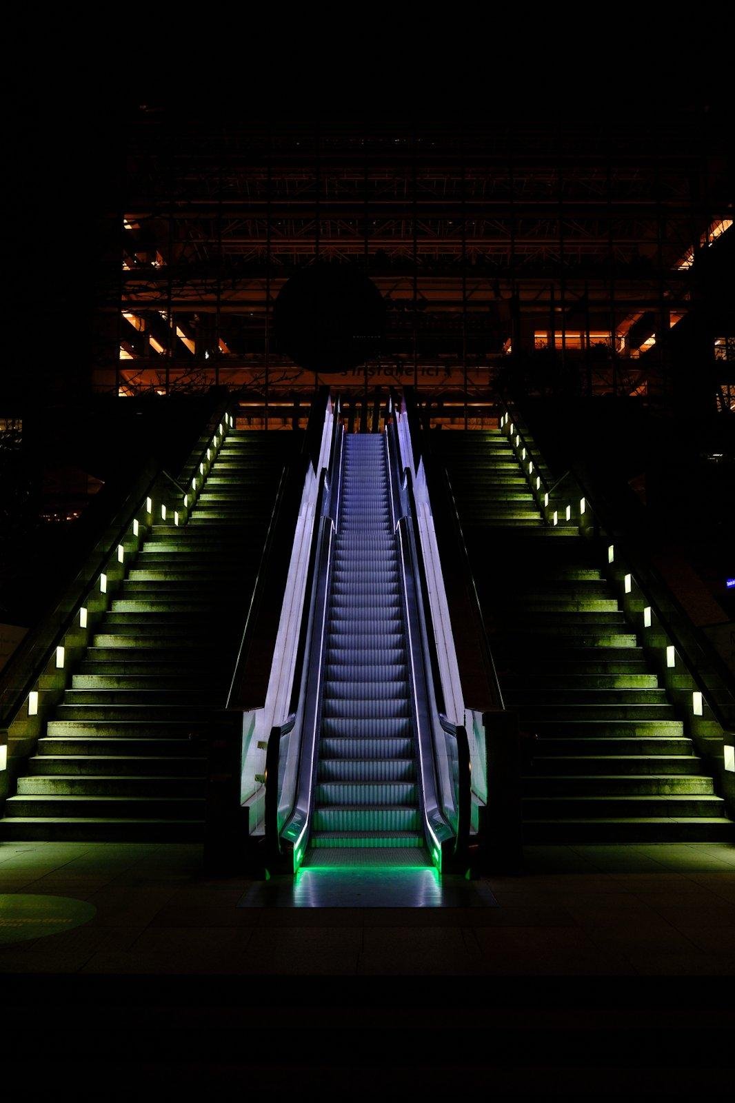 Photo format portrait de nuit en couleur, montrant un escalator éclairé par un ruban de LED tout le long, et un rayon de lumière verte en marque le commencement. De chaque côté se trouvent des escaliers éclairés par des carré de lumière disposés dans le mur à interval régulier, donnant une teinte verdâtre sur la moitié de la largeur des escaliers. Il y a un palier discernable à mi hauteur, cassant la monotonie des lignes. Au sommet on distingue un bâtiment dont une faible lumière orange émane.
