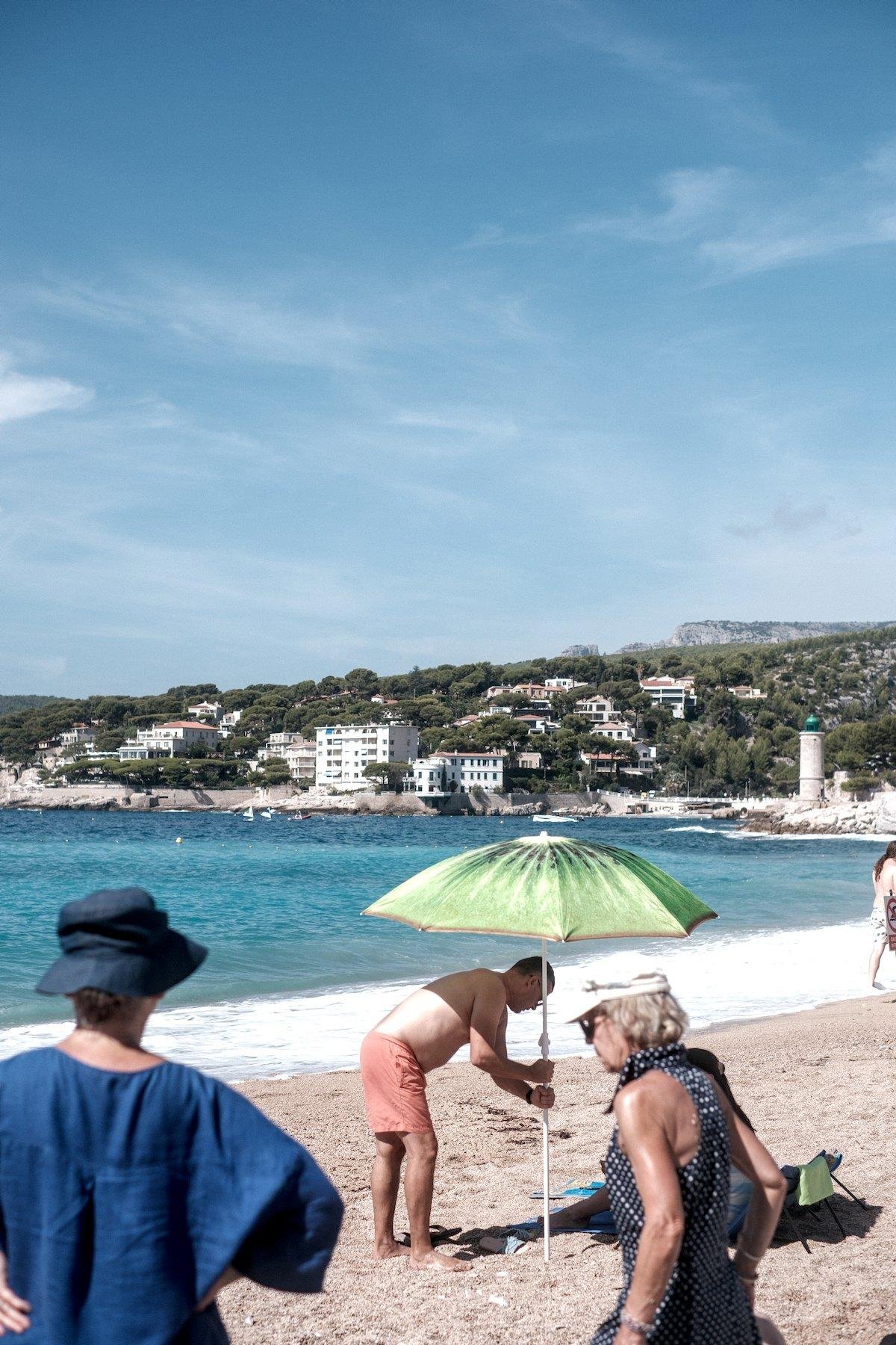 Photo verticale. Sur une plage de sable, au premier plan, 2 dames, celle de gauche de dos, tout de bleu, chapeau inclus, à droite en haut sans manche, casquette blanche, allant vers la gauche. Au second plan, un homme en short plante un parasol dont le motif représente la pulpe verte à pépin noir d’un kiwi. En arrière plan, la mer et encore derrière, un bout de ville mélangé à de la verdure, sous un ciel bleu légèrement voilé de nuages épars