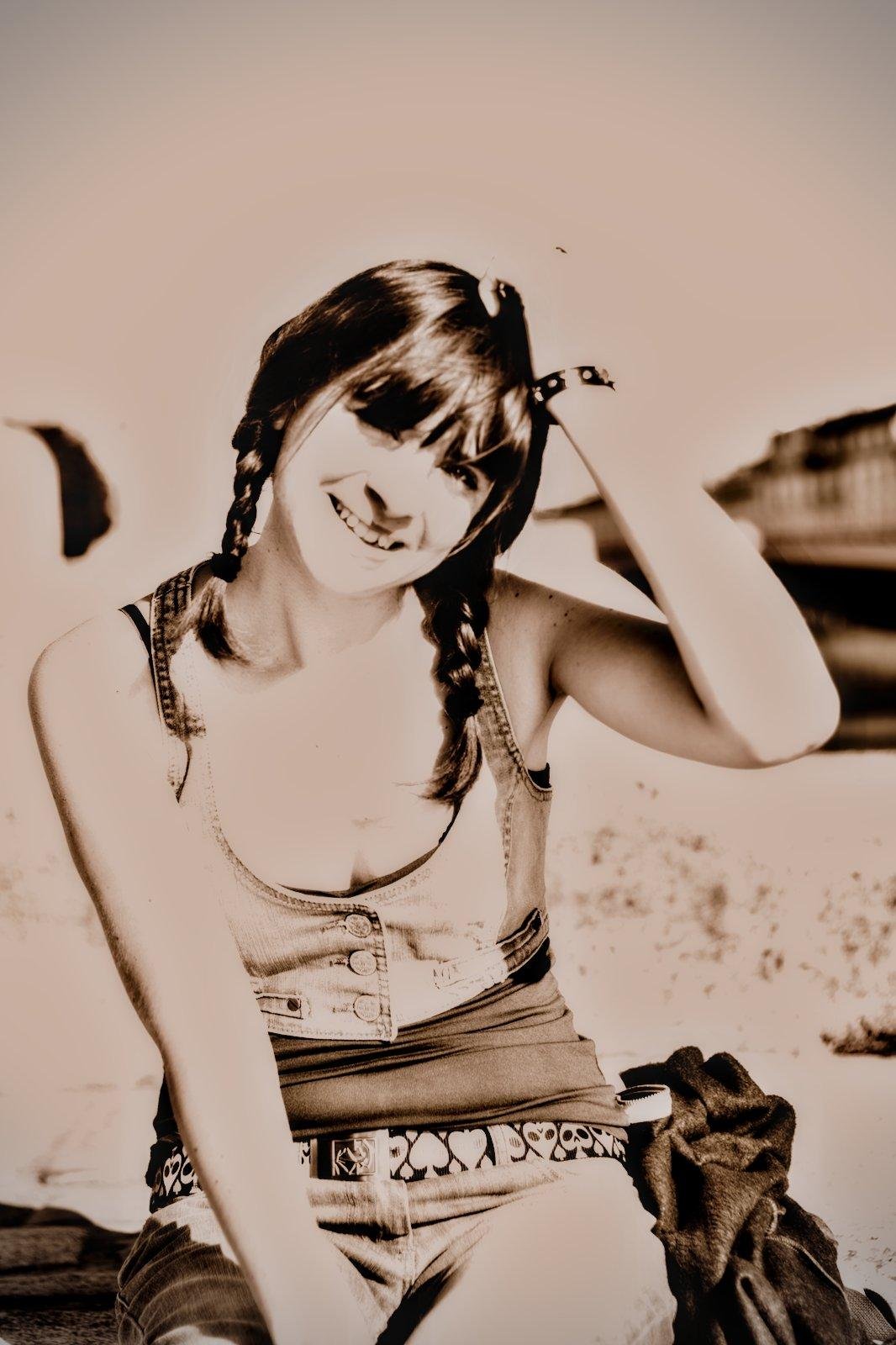 Sepia portrait format photo of a young woman with dark hair tied in 2 braids, scratching her head and smiling