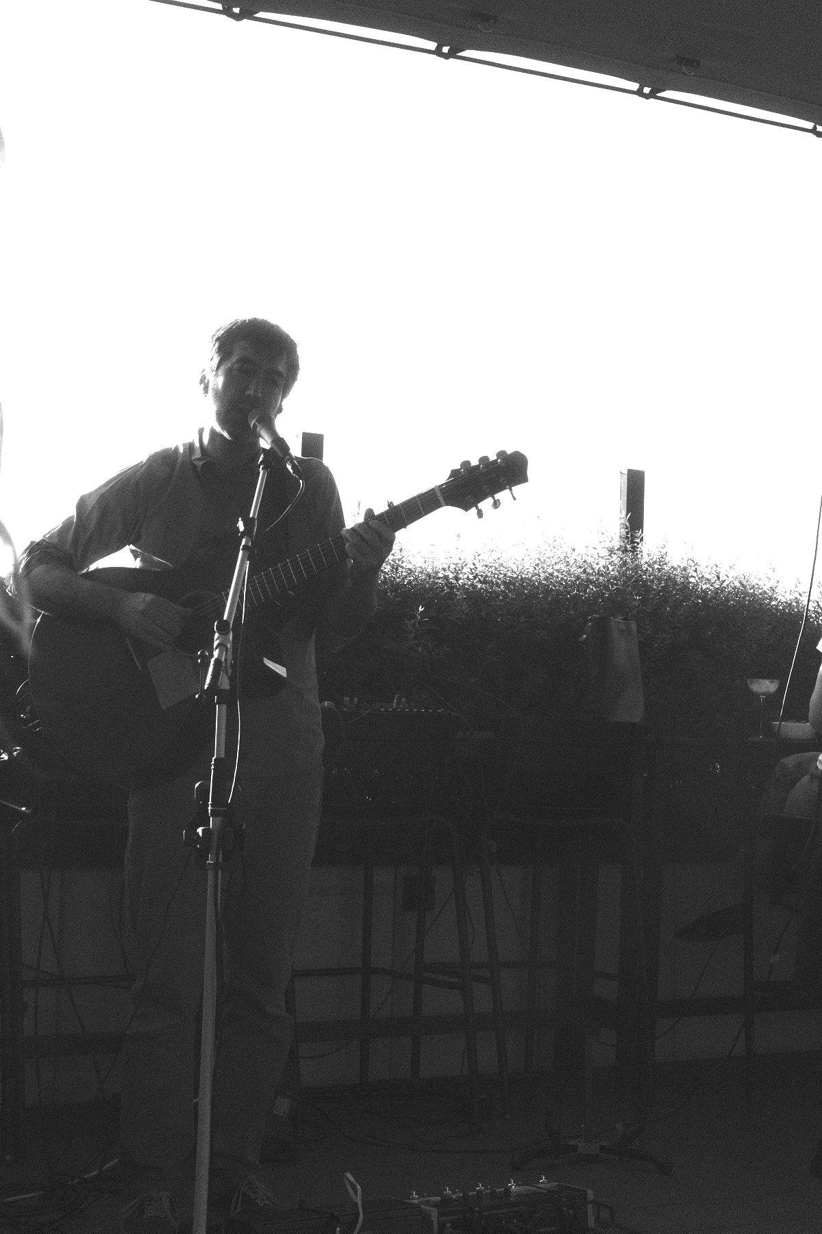 Photo verticale en noir et blanc. Un homme debout chante dans un micro sur pied en jouant de la guitare. Il est devant une haie, sur un ciel très lumineux mettant le musicien en léger contre-jour