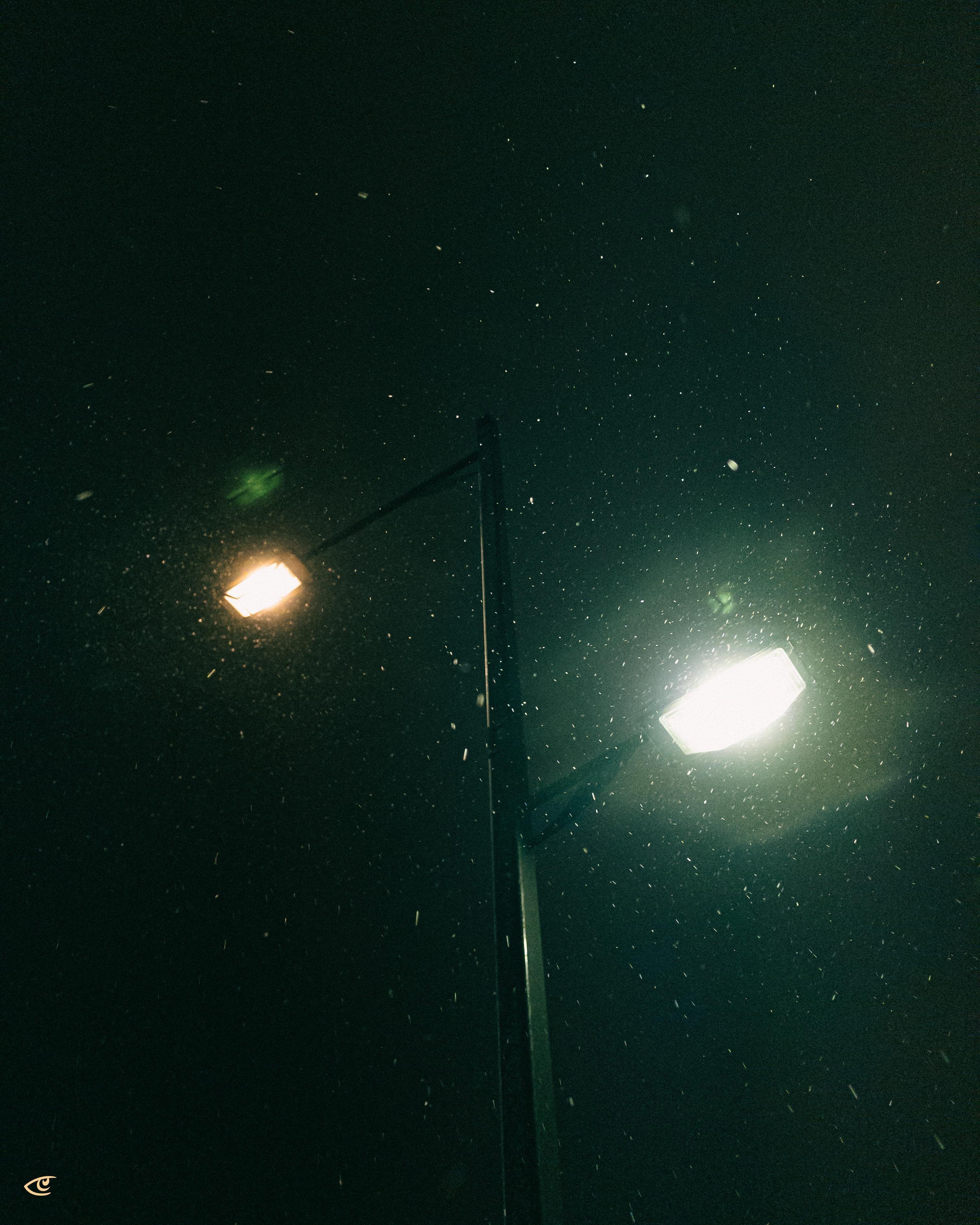 Tall streetlight pole at night with two lamps, one warm yellow and one bright white, illuminating falling snow against a dark sky.