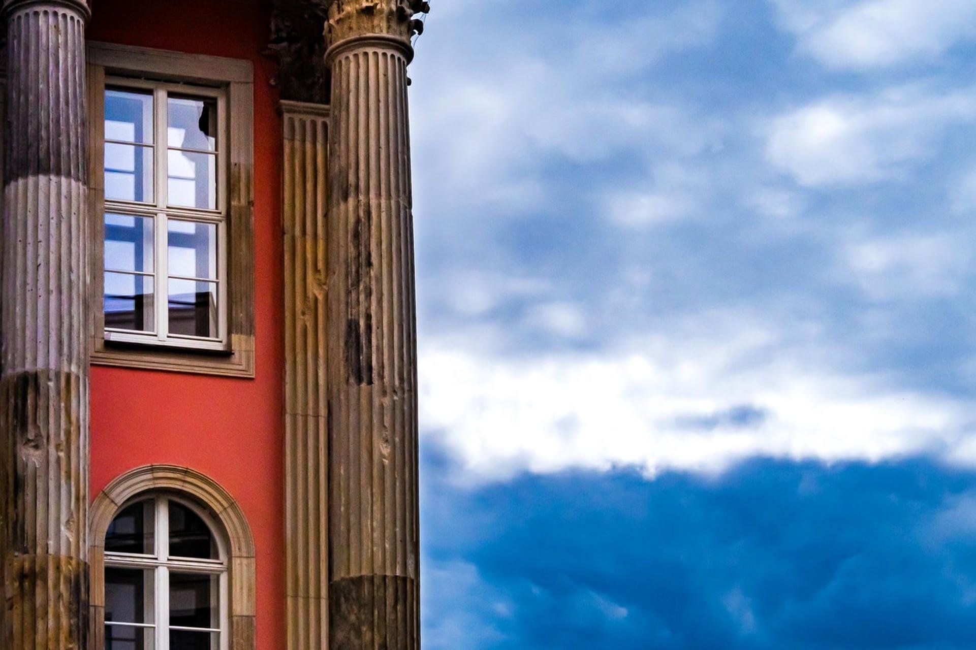 Left side of a red building with two windows framed by tall fluted stone columns, with a cloudy blue-gray sky filling the right side.