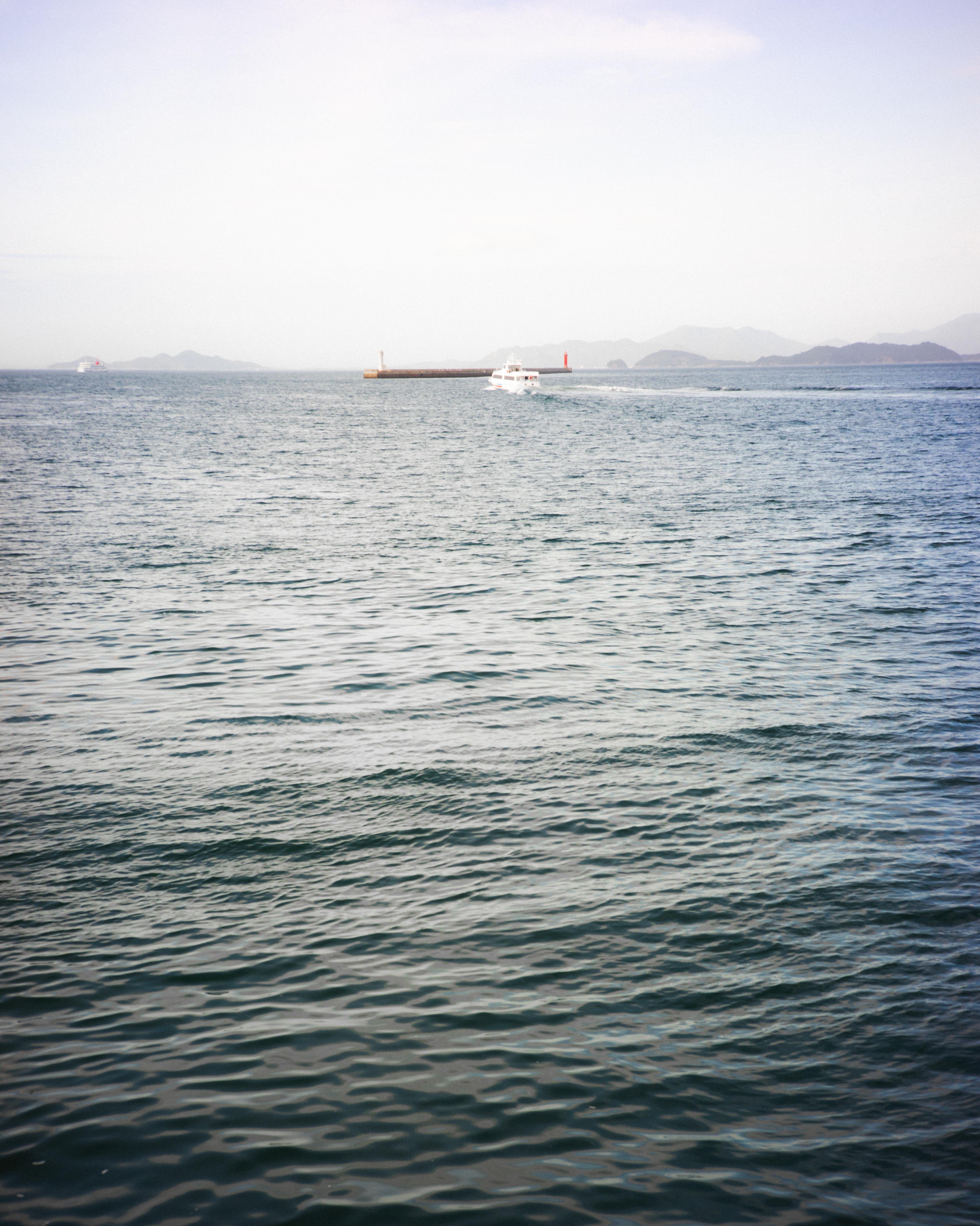 Calm ocean with rippling water in the foreground, a small white boat with a wake in the midground near a breakwater, and distant hills under a clear sky.