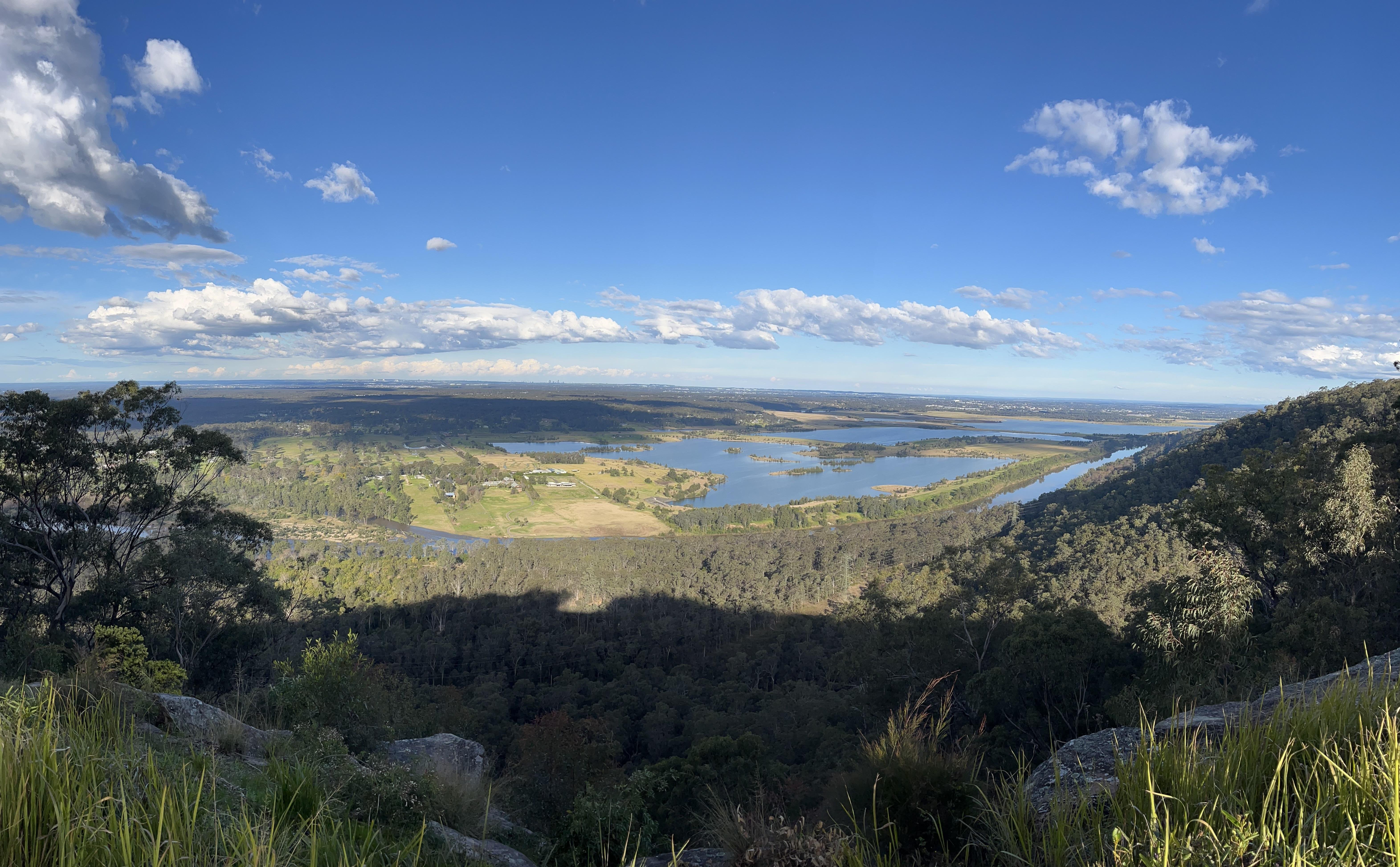 Panoramic landscape featuring a broad view of a valley with a large, reflective lake in the center. The foreground includes grassy areas with rocks and shrubs. Dense forest covers the midground, leading to open fields and patches of greenery surrounding the lake. A horizon of distant rolling hills under a clear blue sky with scattered clouds.