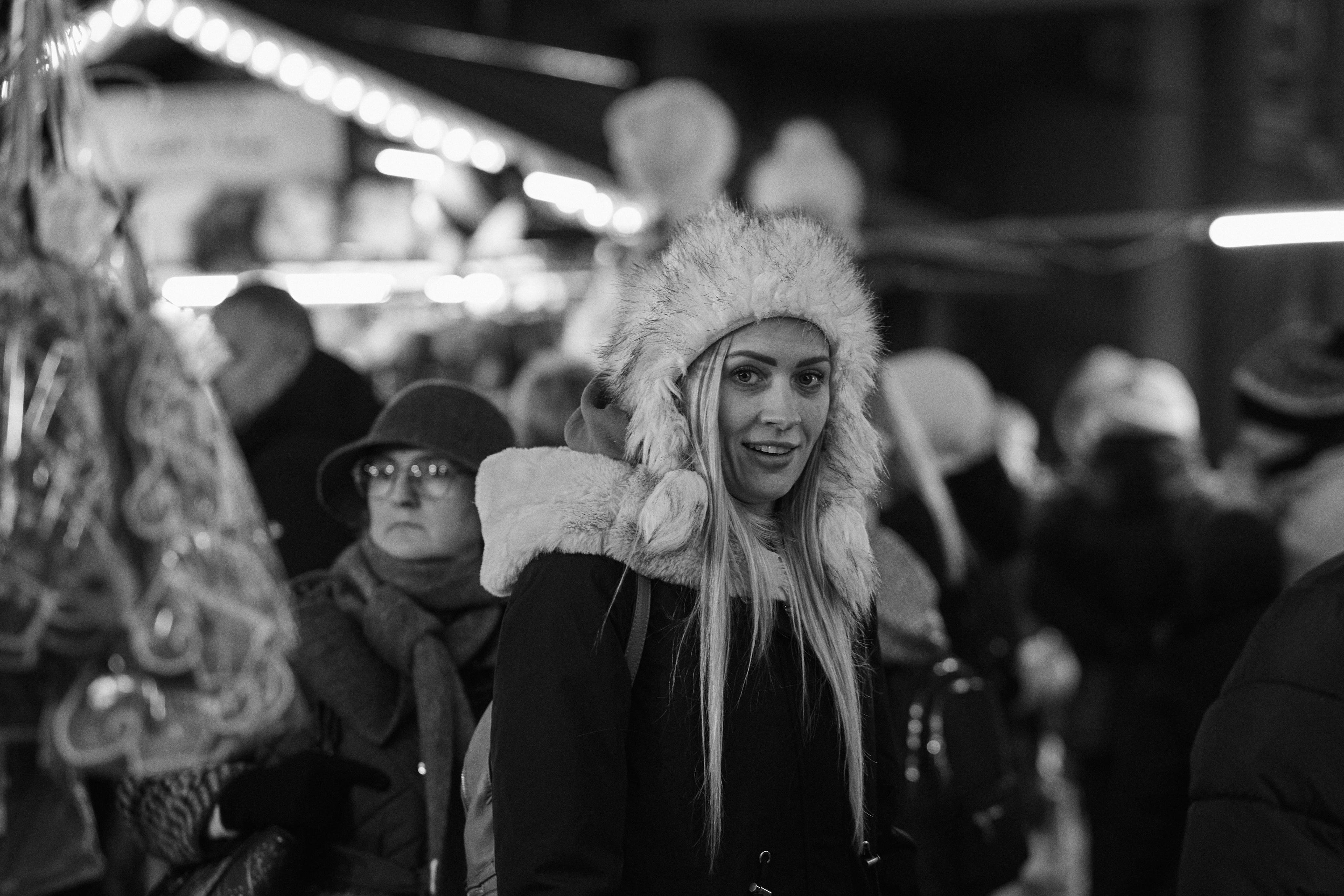 The image shows a black-and-white photo of a woman in a crowd. She's wearing a furry hat with earflaps and a coat with a fur hood. The background is busy, likely at a market or festive event with bright lights and other people bundled in winter clothing.