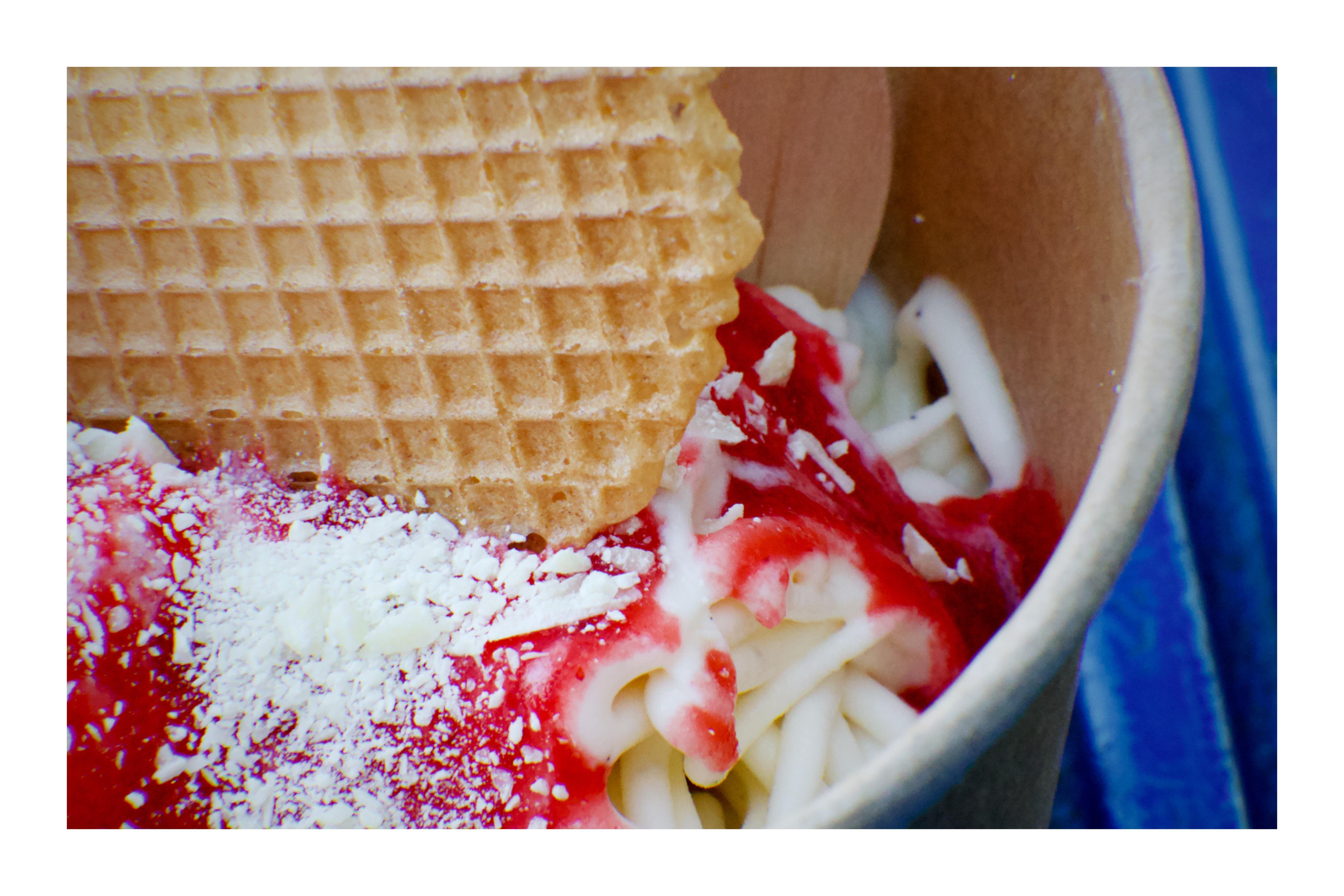 This image shows a close-up of a serving of spaghetti ice cream. It appears to have vanilla ice cream pressed to look like spaghetti noodles, topped with a red sauce resembling strawberry sauce, grated white chocolate simulating parmesan cheese, and a wafer.