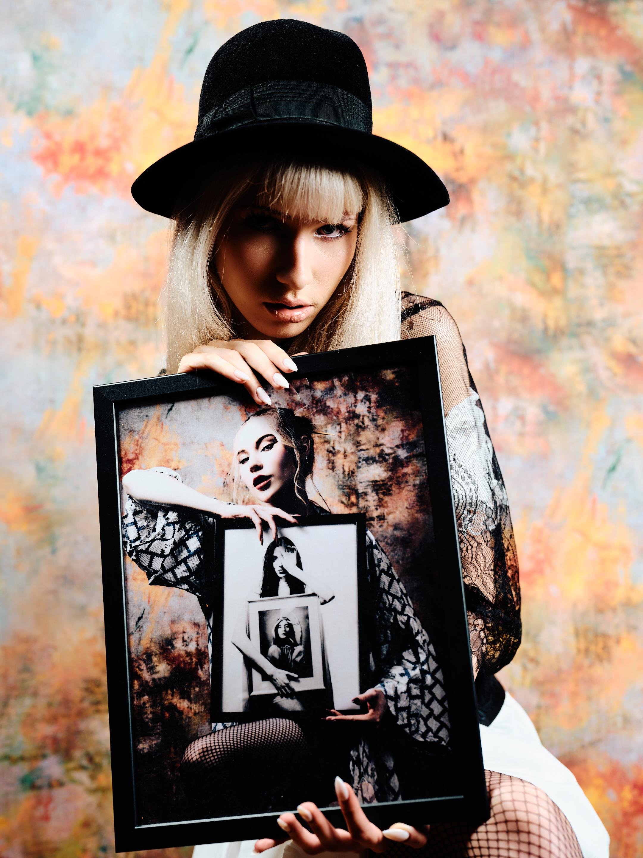 Blonde woman wearing a wide-brim black hat and lace sleeves holds a large framed photo in front of her torso; the photo shows her holding another framed photo, creating a repeating image effect, against a mottled orange, blue, and beige background.
