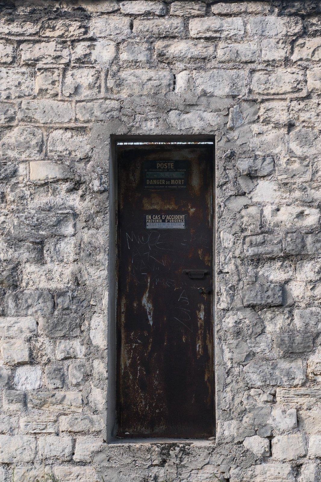 Photo verticale. Une porte étroite en métal et toute rouillée encastrée dans un mur en vieille pierres. Sur une plaque, on peut lire entre autre, «&nbsp;Danger de mort&nbsp;»