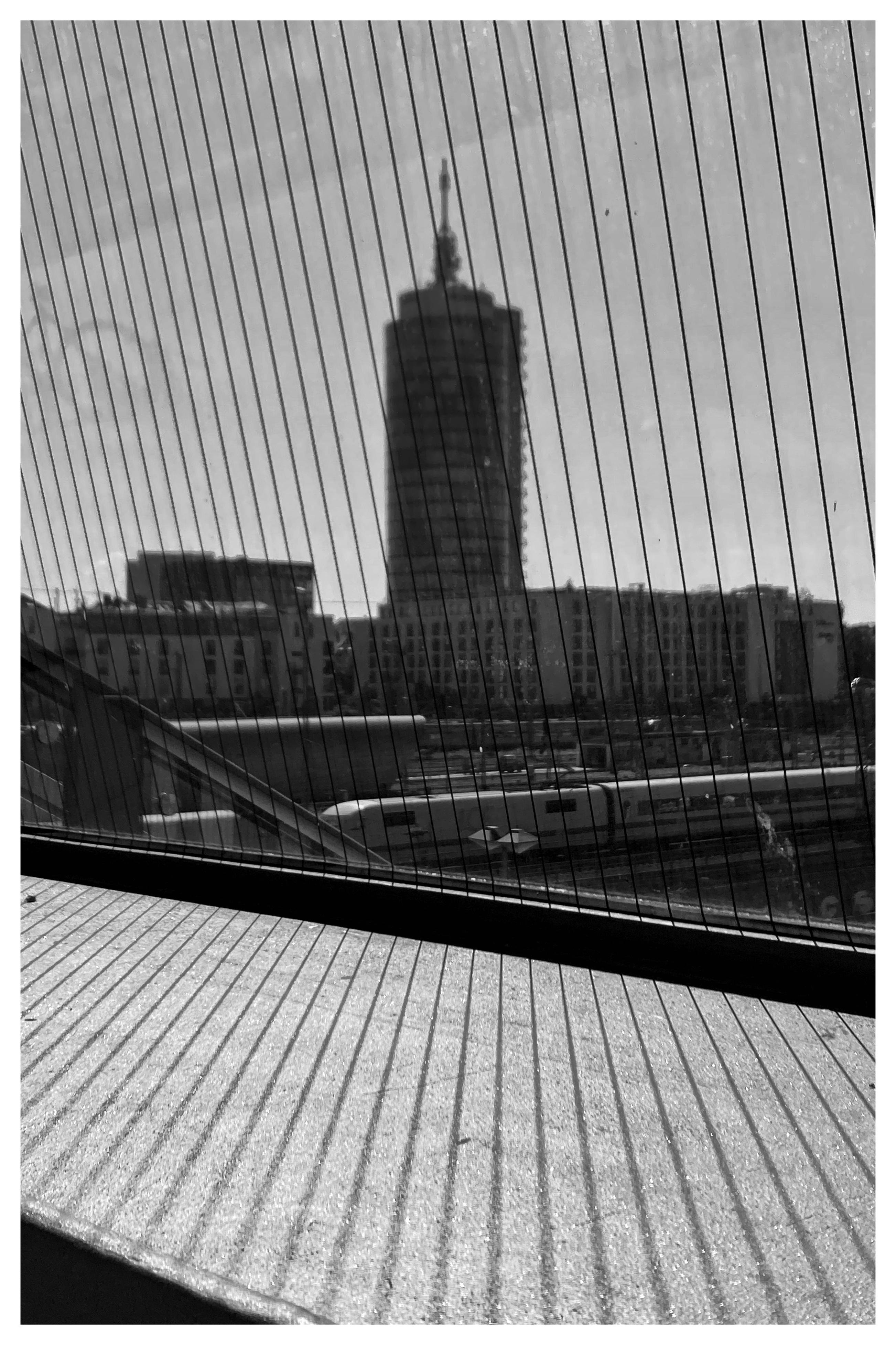 Black and white image of a tall cylindrical building under construction, viewed through vertical lines of a window. The building is flanked by lower, rectangular structures. In the foreground, diagonal shadows from the window lines cast onto a flat surface. Sky is clear with minimal cloud cover.